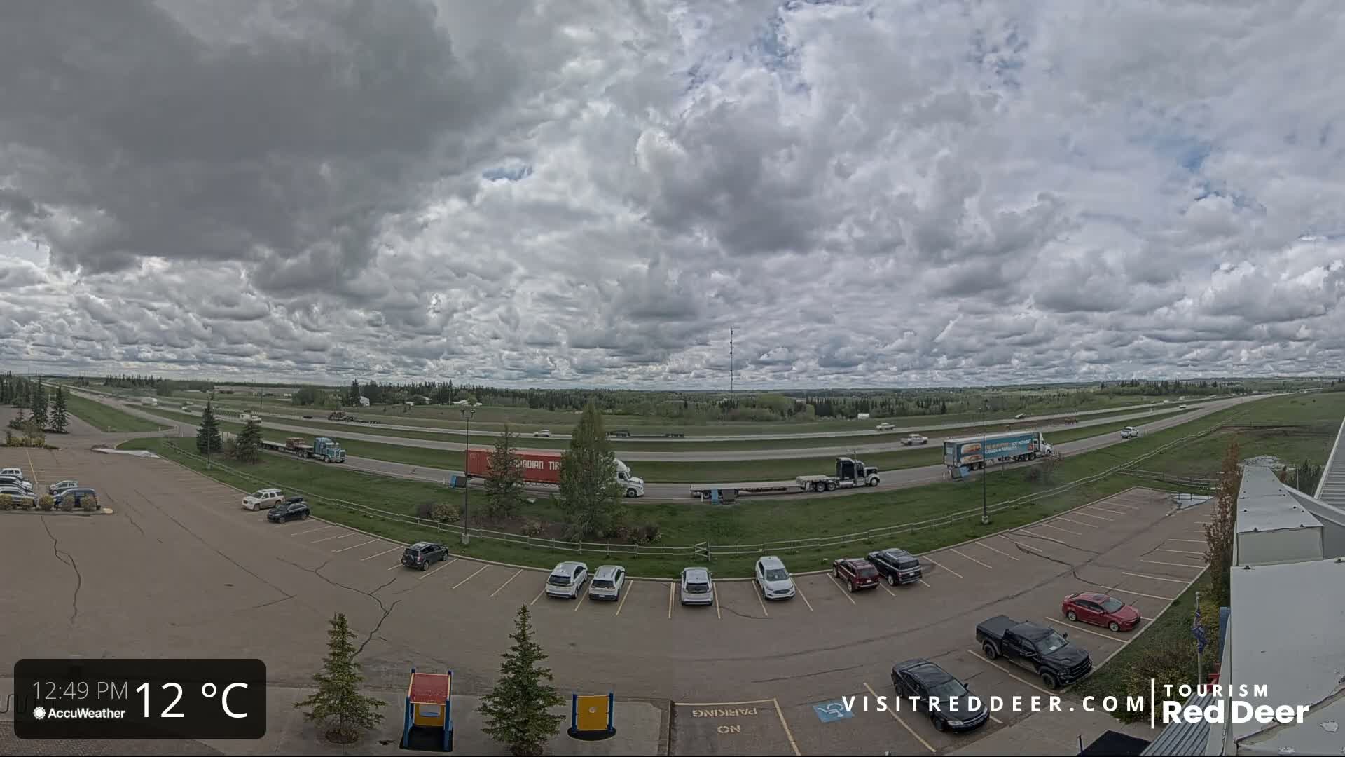 A partly cloudy sky over a highway and parking lot with several cars and semi-trucks.