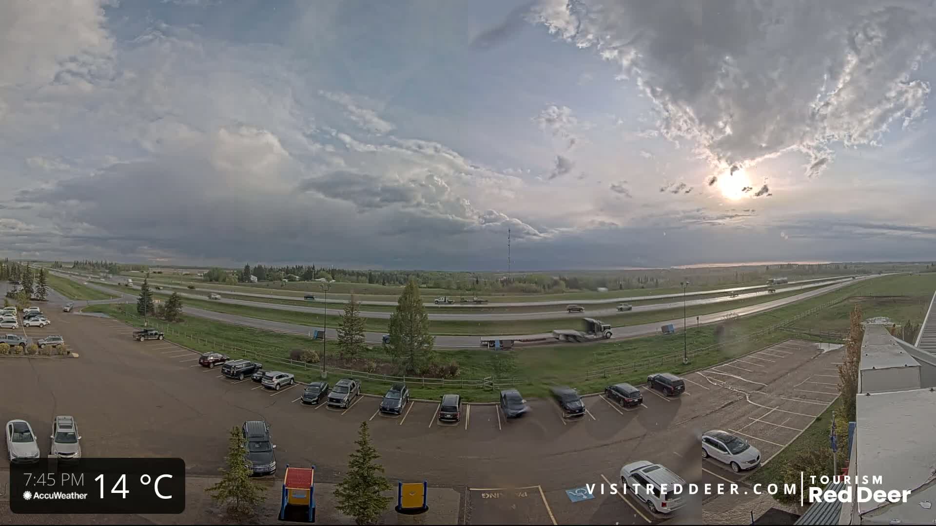 A partly cloudy sky with the sun shining through illuminates a highway with several cars and trucks driving, and a parking lot with numerous vehicles parked next to a building.