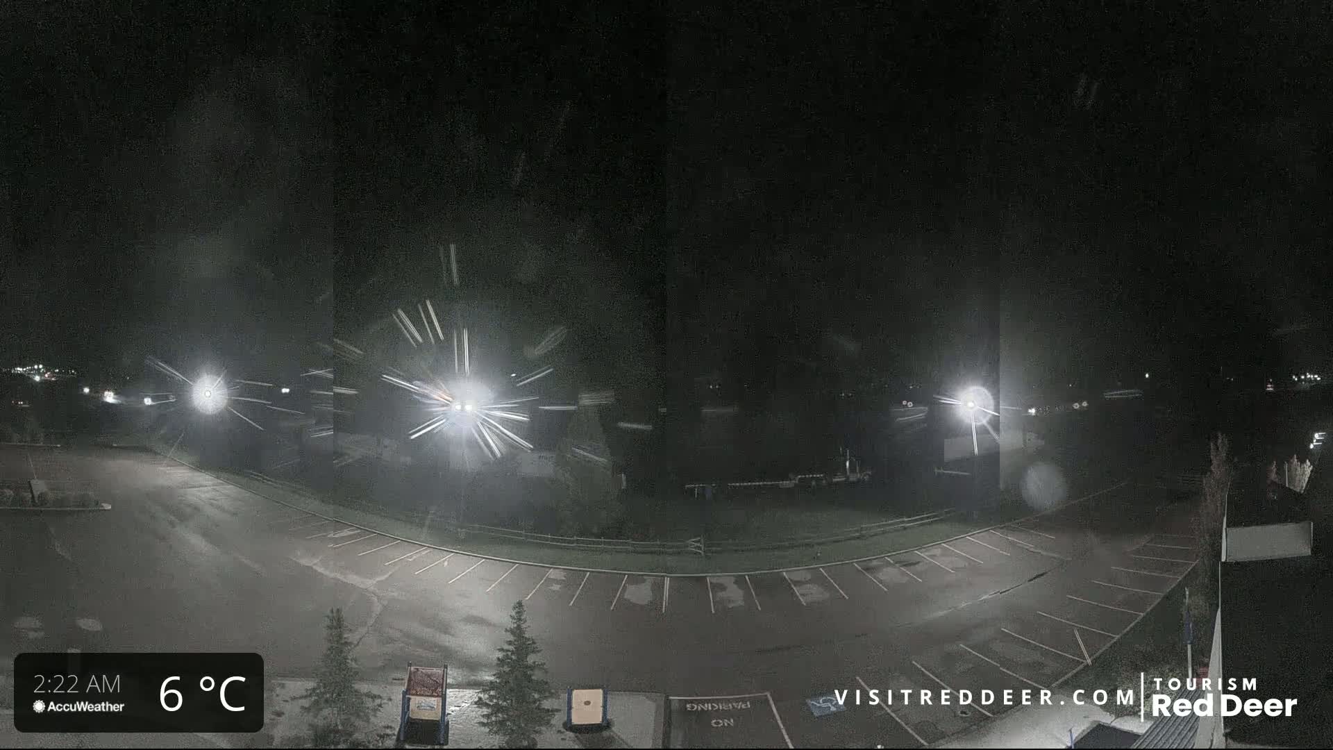 A nighttime view of a mostly empty parking lot with light snow falling, illuminated by several bright lights.