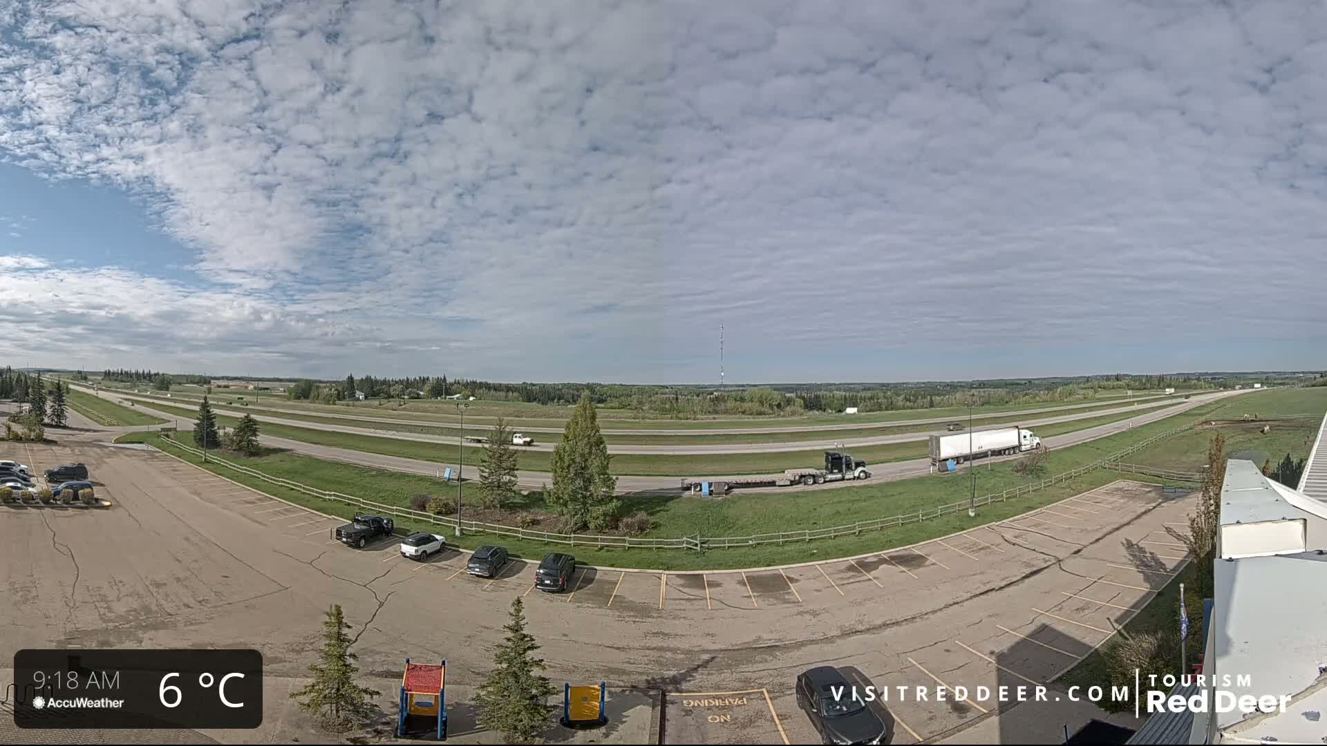 A partly cloudy sky over a highway with several vehicles in a parking lot and a few vehicles traveling on the road, the temperature is 6°C.