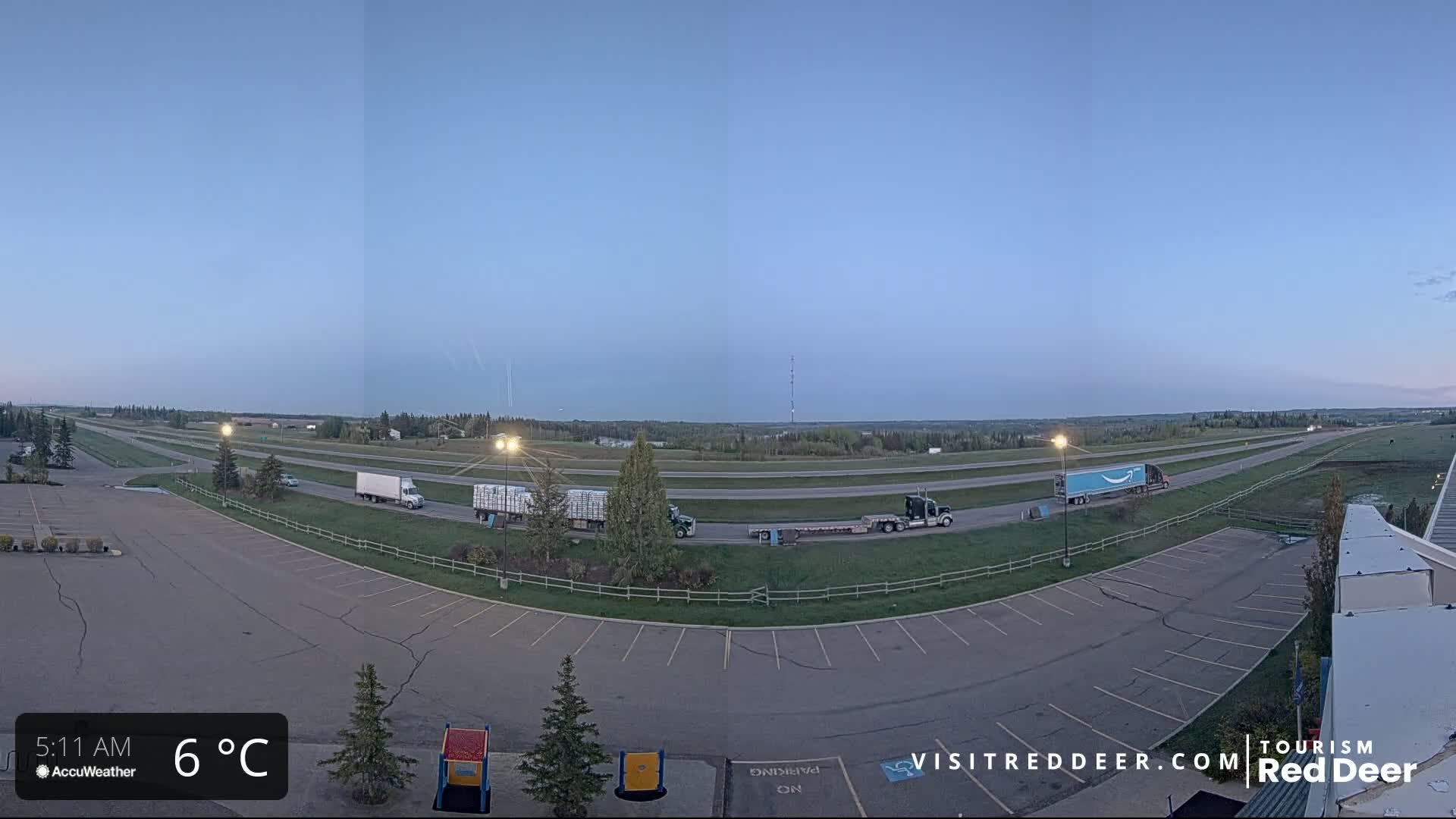 A wide shot of a highway with several semi-trucks driving on it at dawn in 6°C weather, with an empty parking lot visible in the foreground.