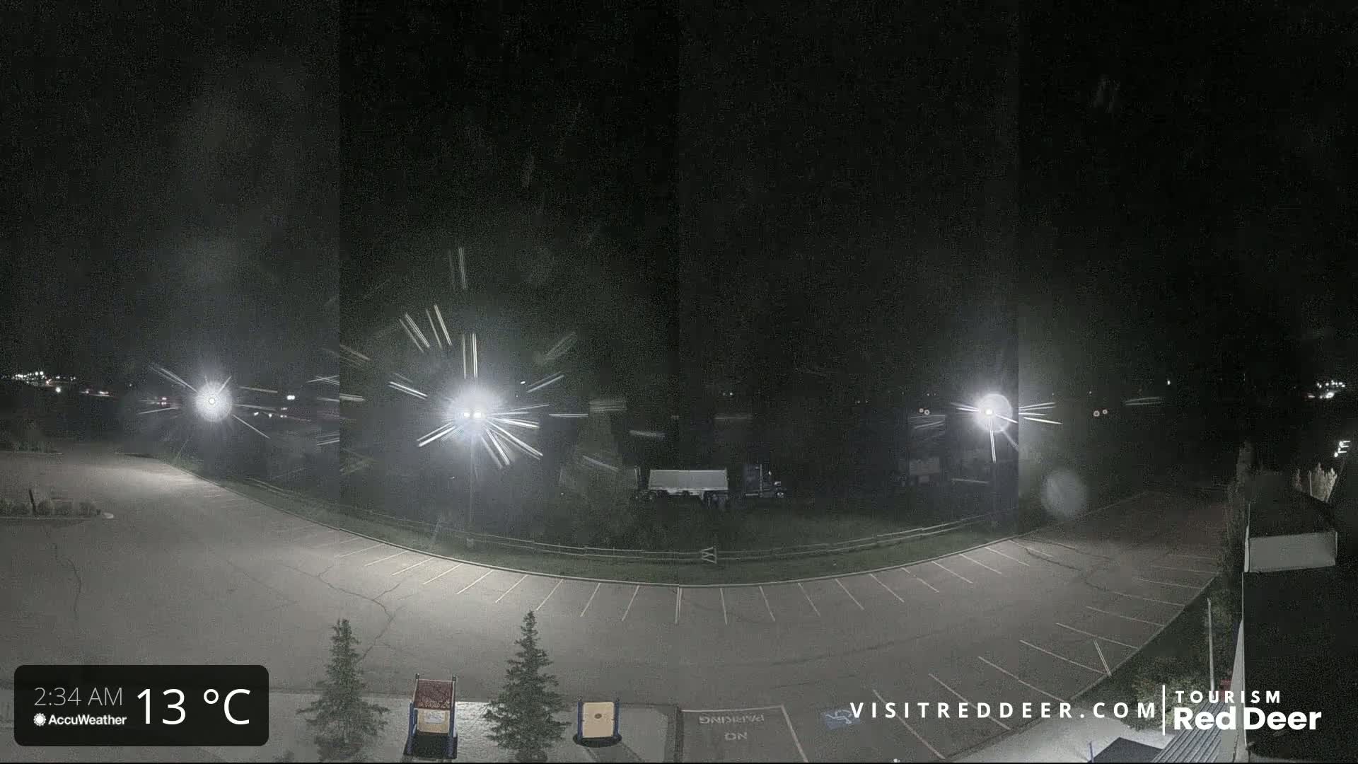 A nighttime view of an empty parking lot with several bright lights and some light snow or rain falling.
