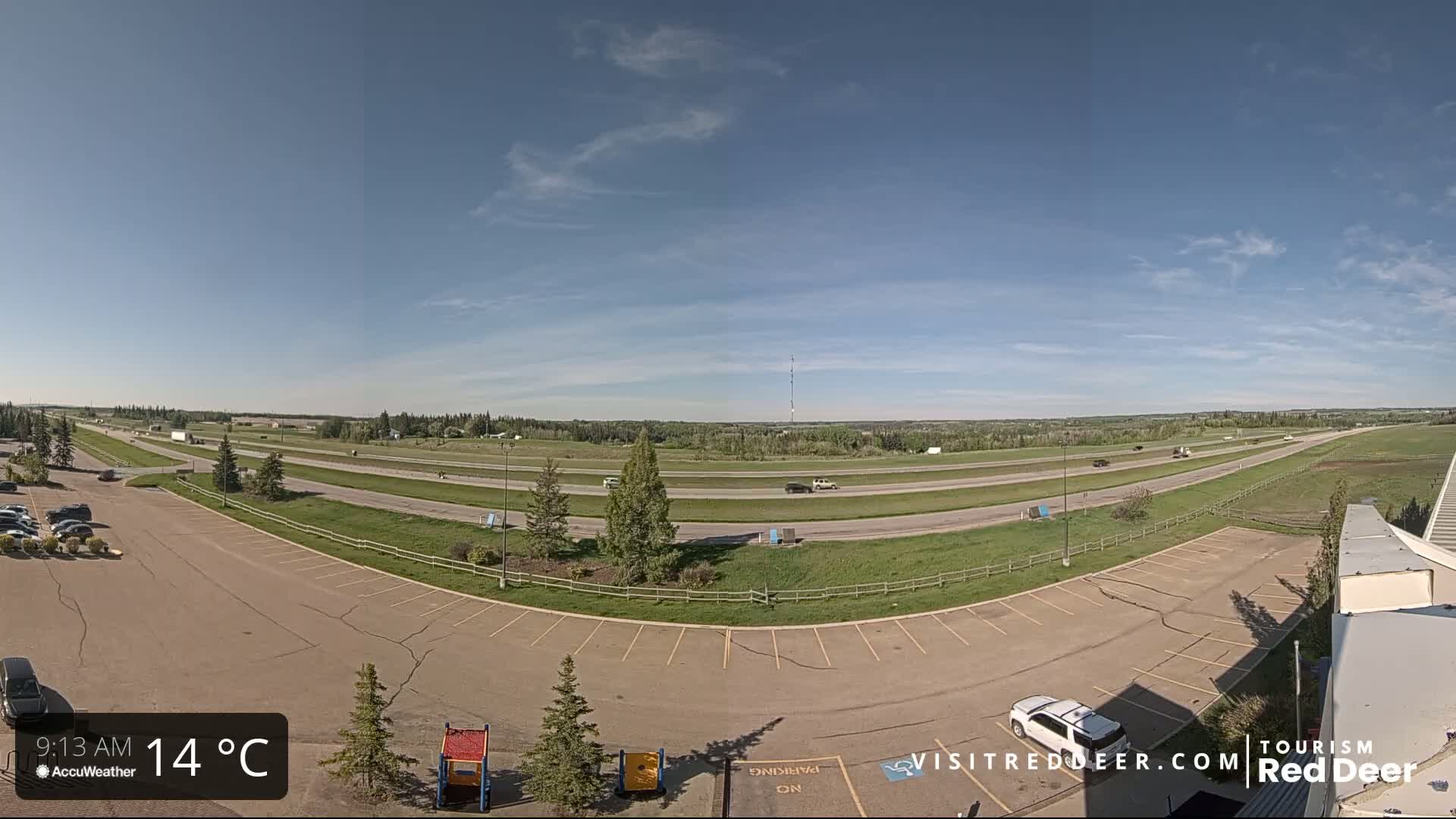 A mostly sunny day shows a highway with light traffic, a large empty parking lot, and a grassy area beyond the road.
