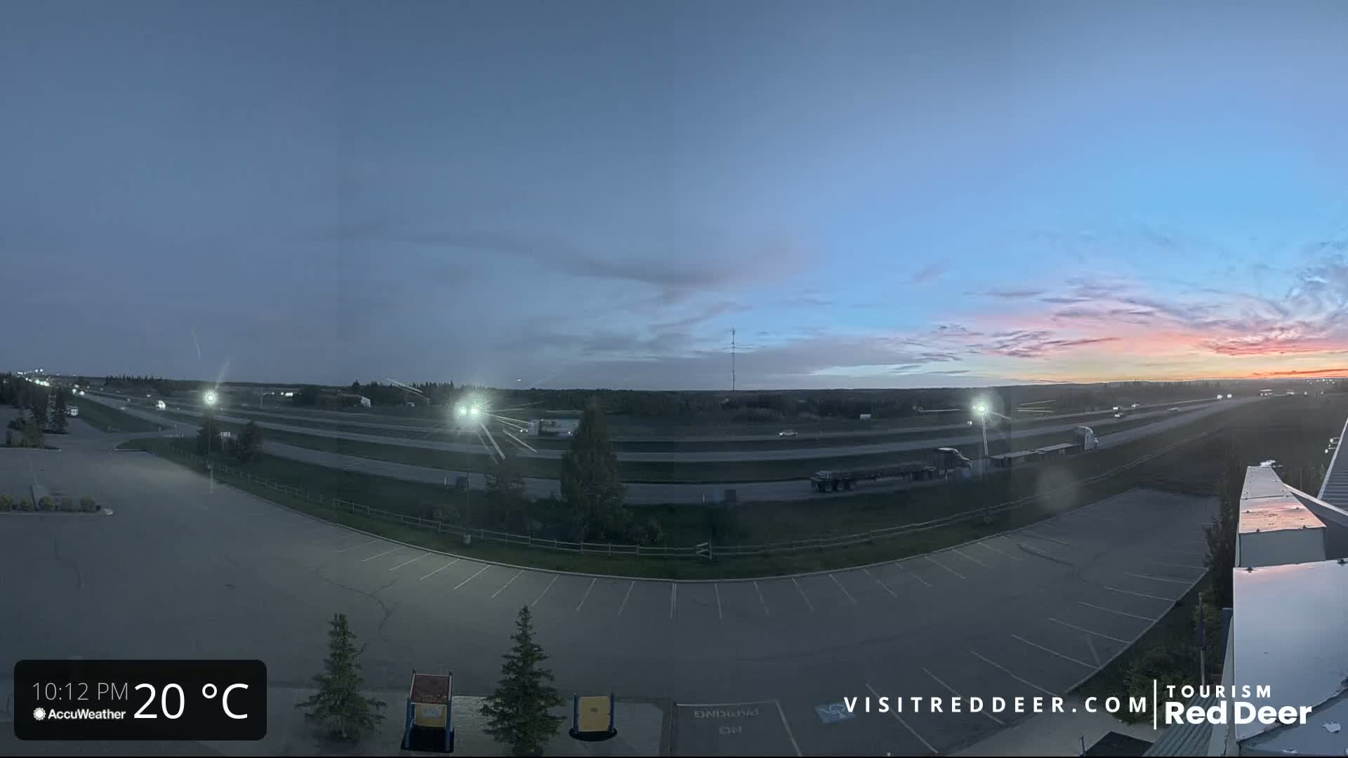 A twilight scene shows a highway with some traffic, an empty parking lot, and a pinkish sunset in the distance under a mostly clear sky.
