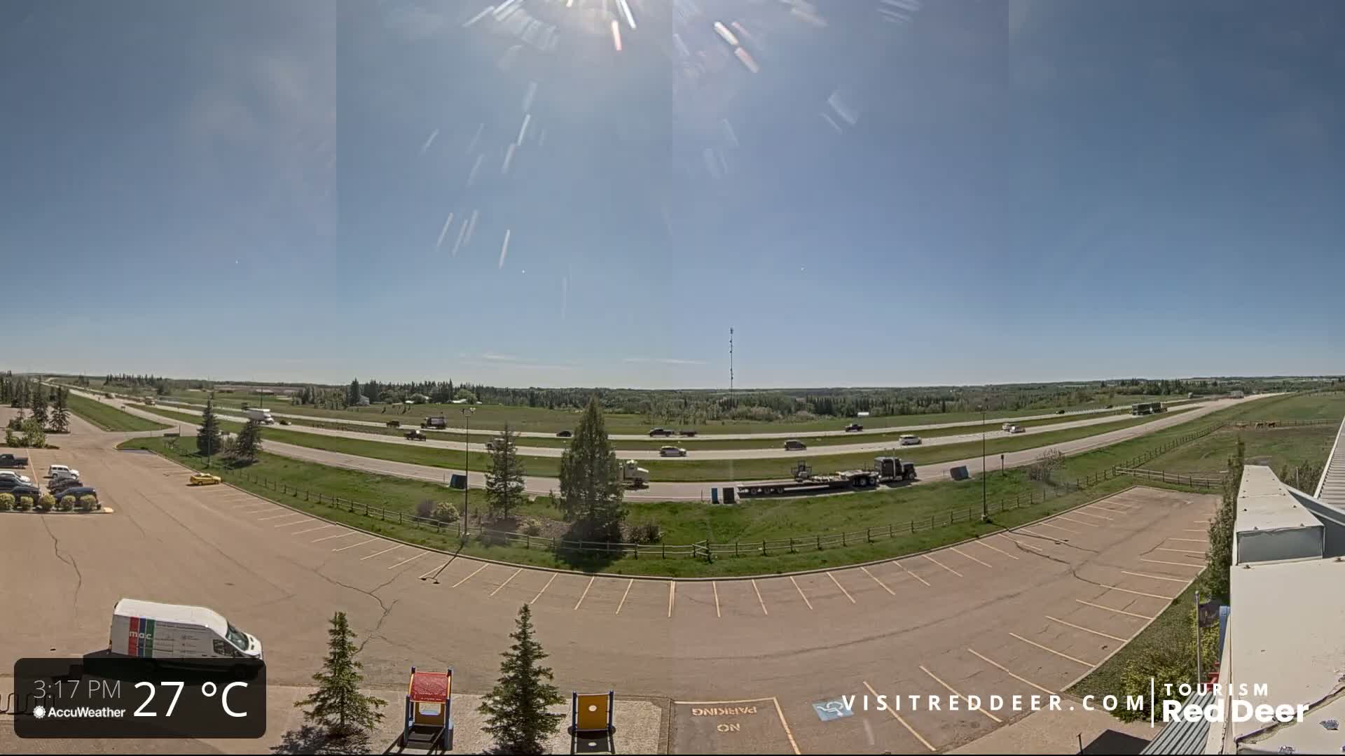 A sunny day reveals a highway with moderate traffic, a large parking lot, and green fields in the background.
