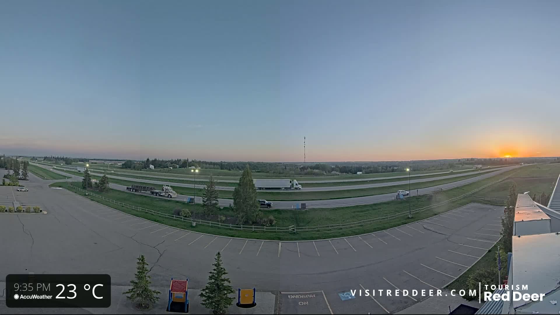 An empty parking lot overlooks a highway with several vehicles, including semi-trucks, during a sunset with clear skies and a temperature of 23°C.