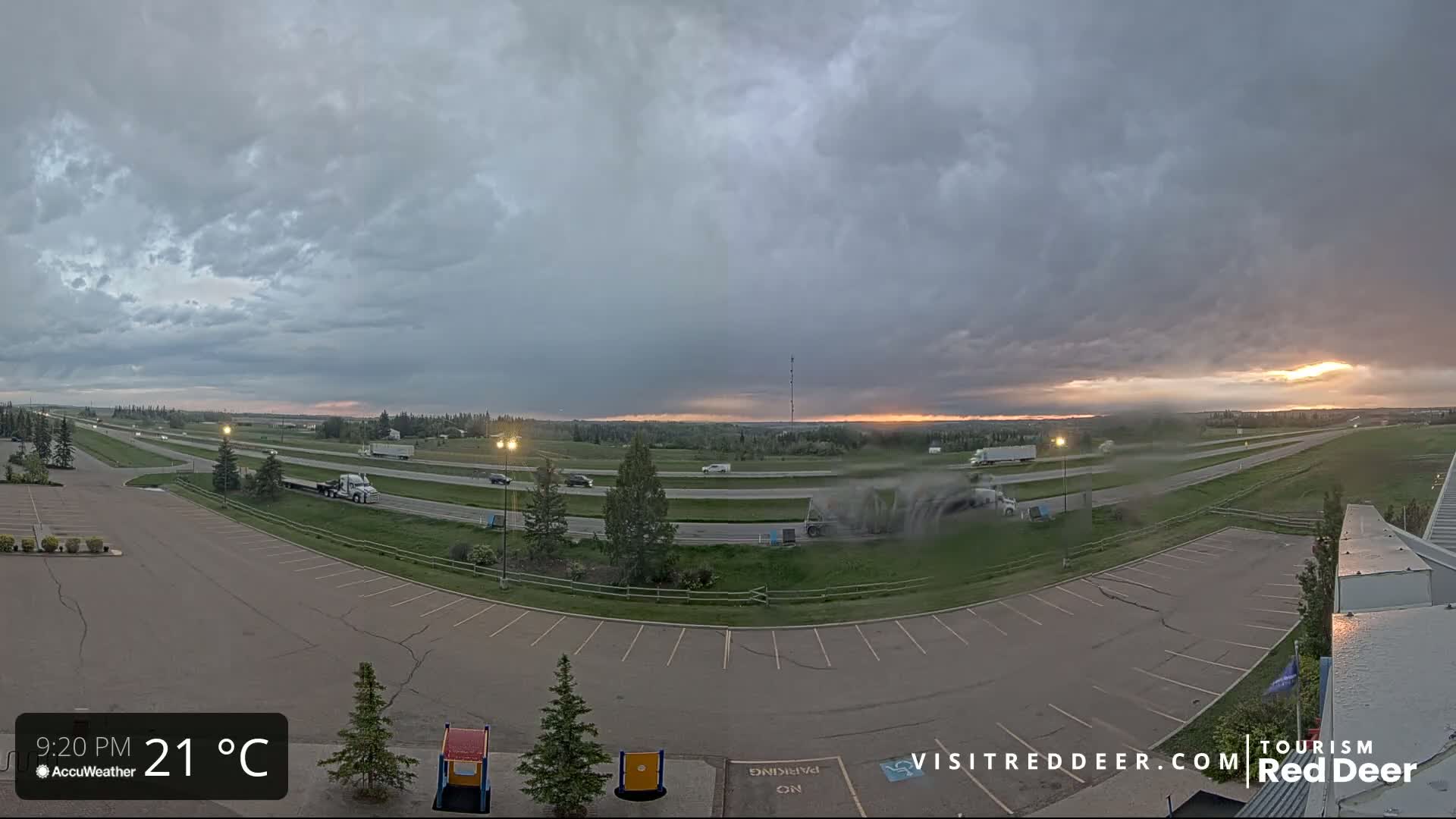 A highway with several vehicles is visible under a partly cloudy, stormy sky at sunset, with a large empty parking lot in the foreground.