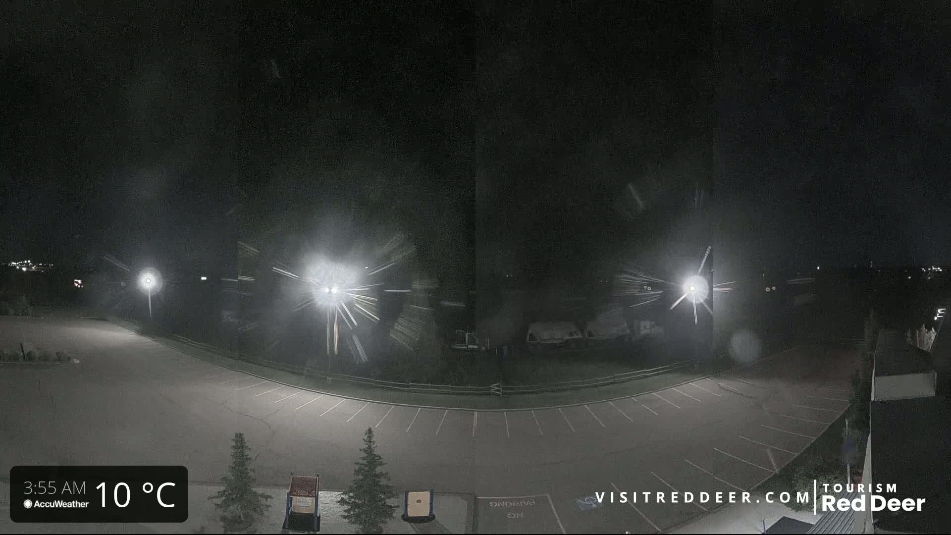 A nighttime view of an empty parking lot with light snow falling, illuminated by several bright lights.
