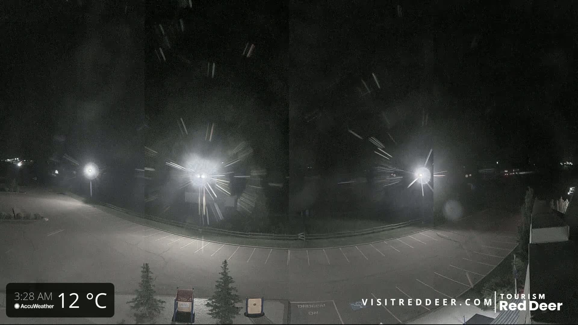 A nighttime, snow-covered parking lot is illuminated by several bright lights, with light snow falling.