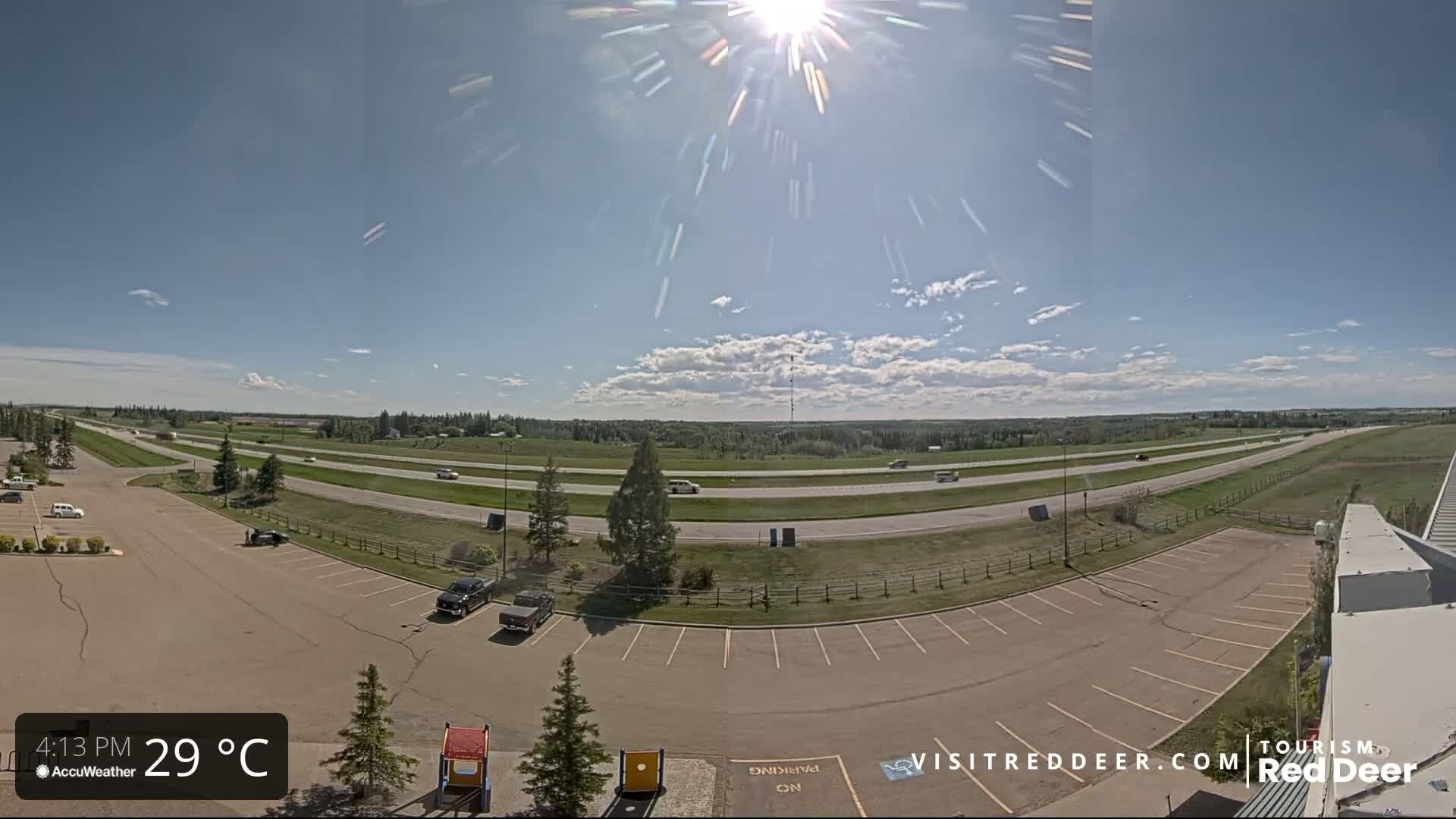 A sunny day with partly cloudy skies shows a highway with sparse traffic next to a mostly empty parking lot.