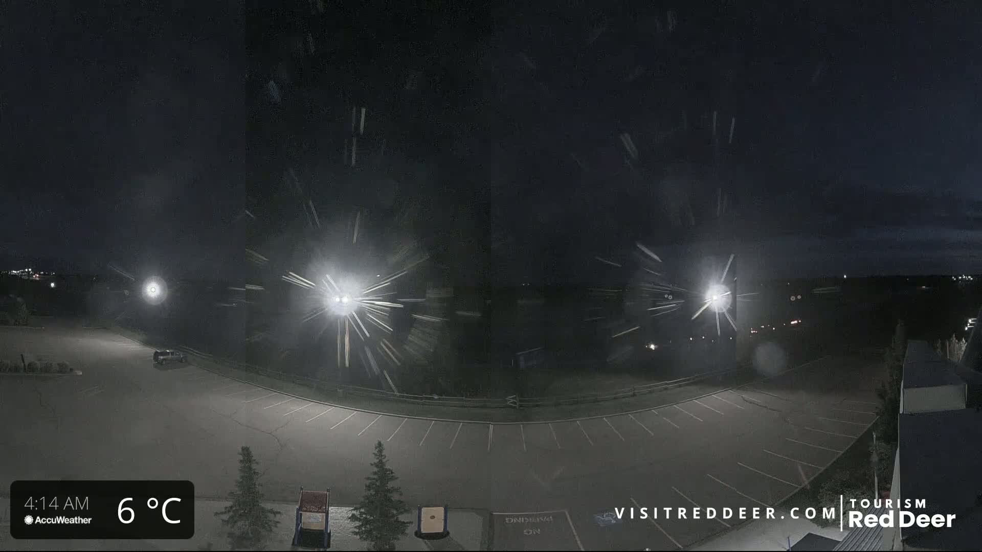 A mostly empty parking lot is seen at night under snowy conditions, illuminated by several bright lights.