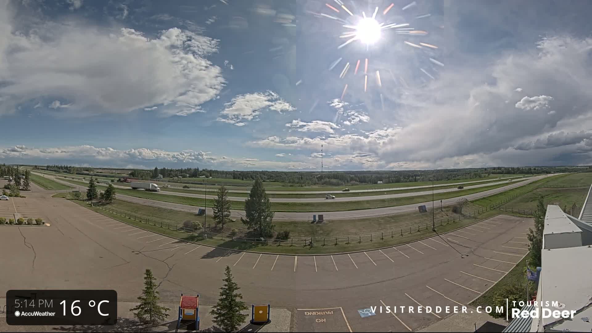 A partly cloudy day with bright sunshine shows a highway with several vehicles and an adjacent empty parking lot.