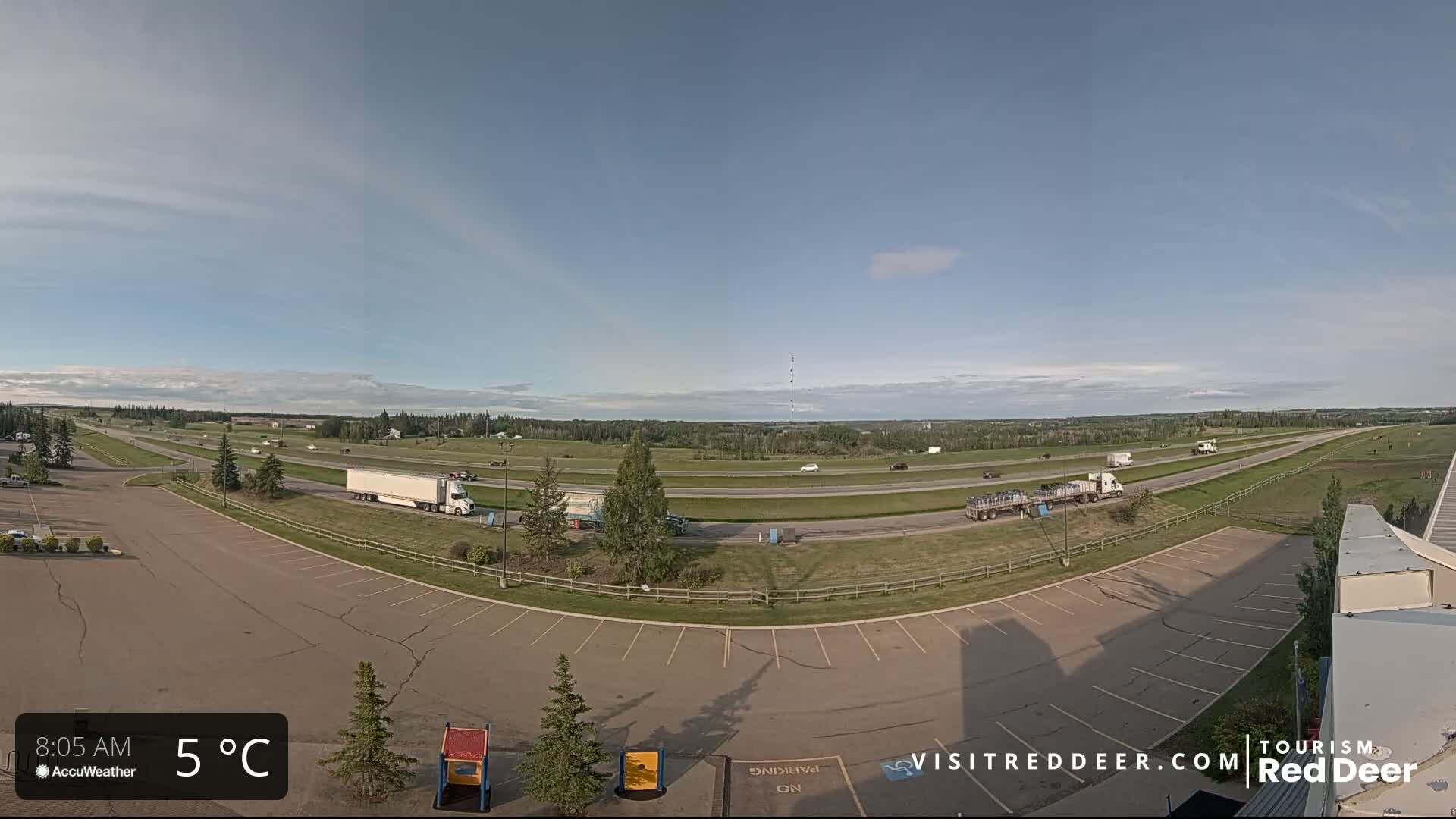 A mostly empty parking lot overlooks a highway with several vehicles and flat, grassy land under a partly cloudy sky.