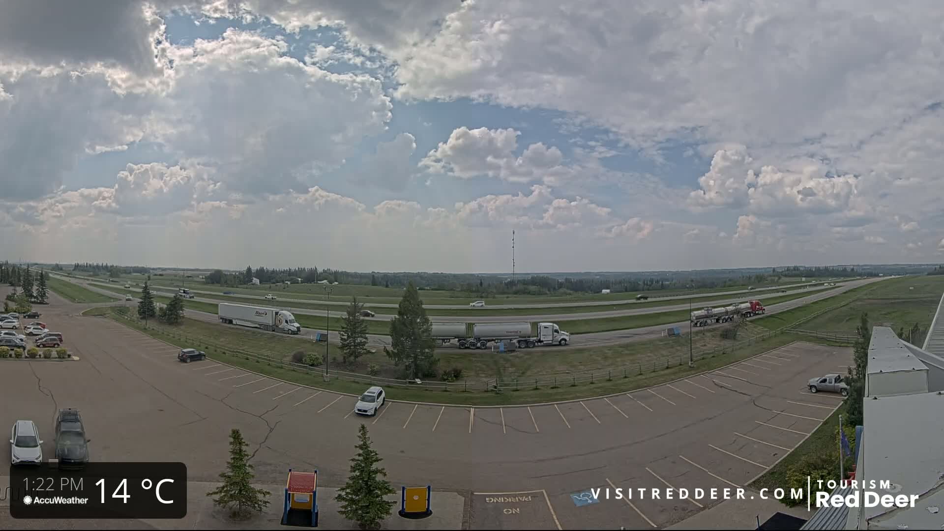 A partly cloudy day with a temperature of 14°C shows a highway with several semi-trucks and cars, as well as a parking lot with cars.