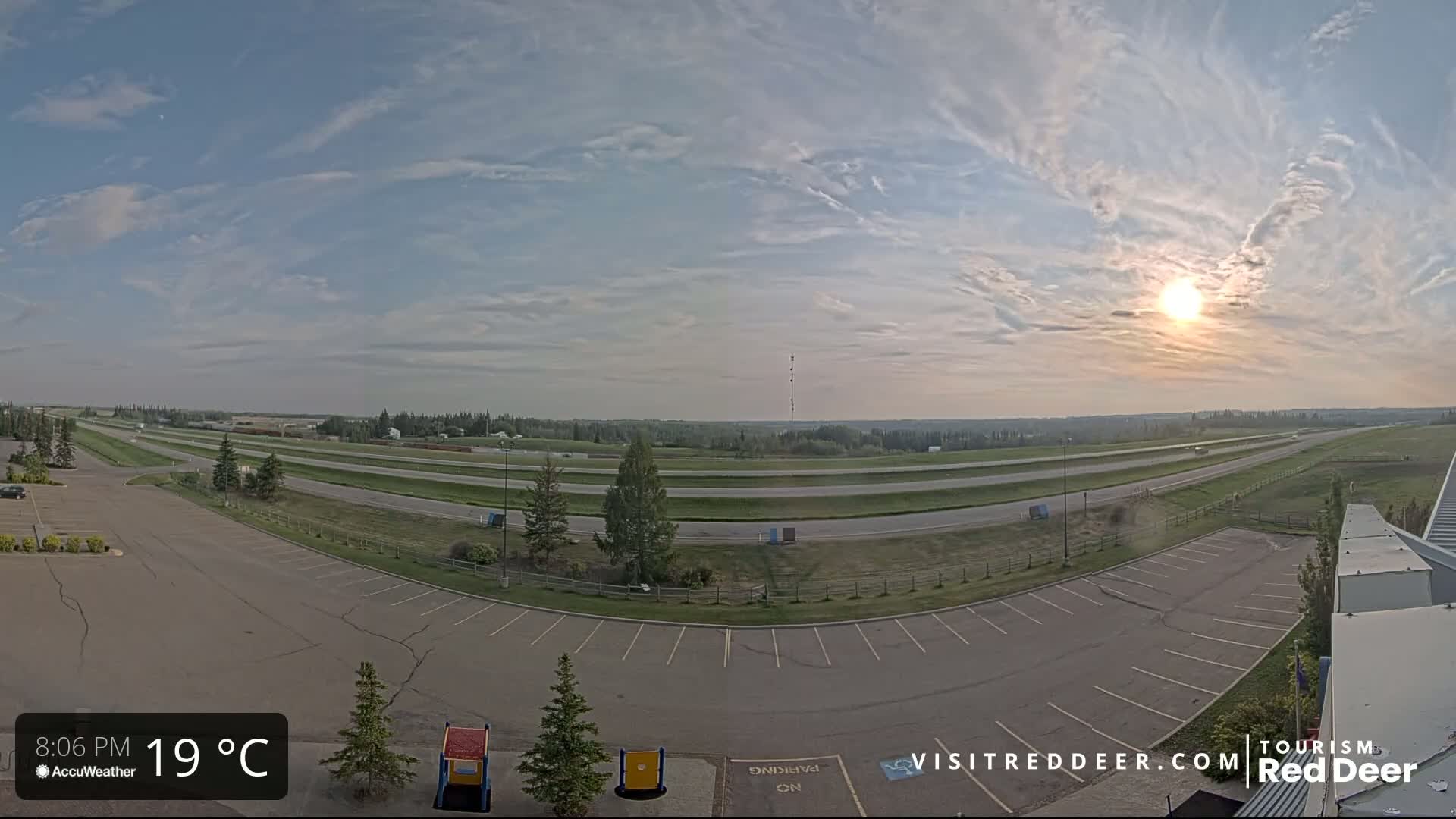 A mostly sunny late afternoon scene shows a wide, multi-lane highway curving through a flat landscape with sparse trees, next to an empty parking lot.