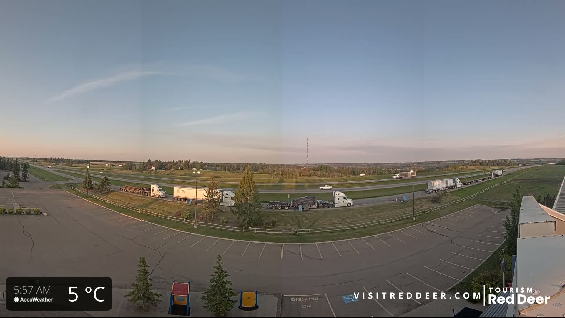 Several semi-trucks are parked in a lot adjacent to a highway under a clear, twilight sky at 5°C.