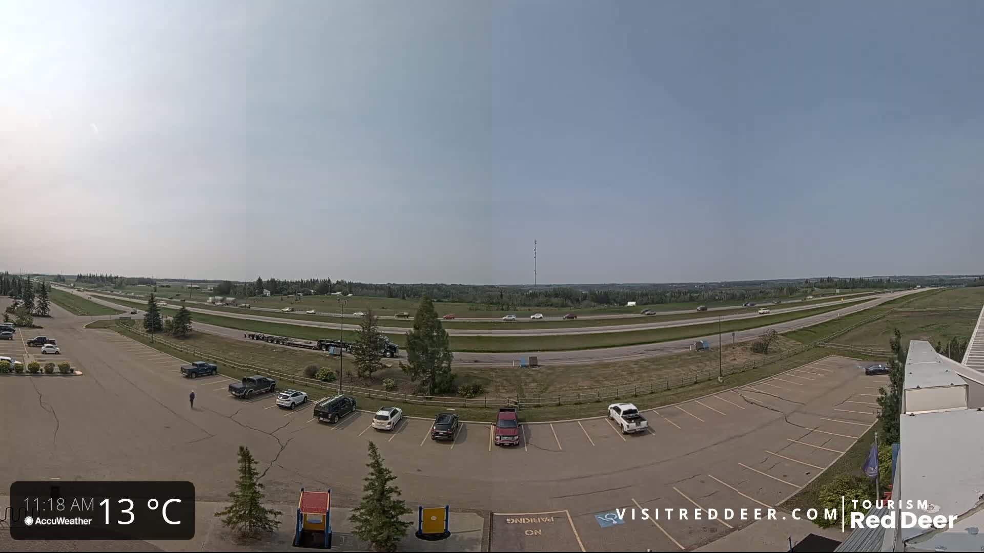 A mostly sunny, 13°C day is shown overlooking a highway with moderate traffic and an adjacent parking lot containing several cars.
