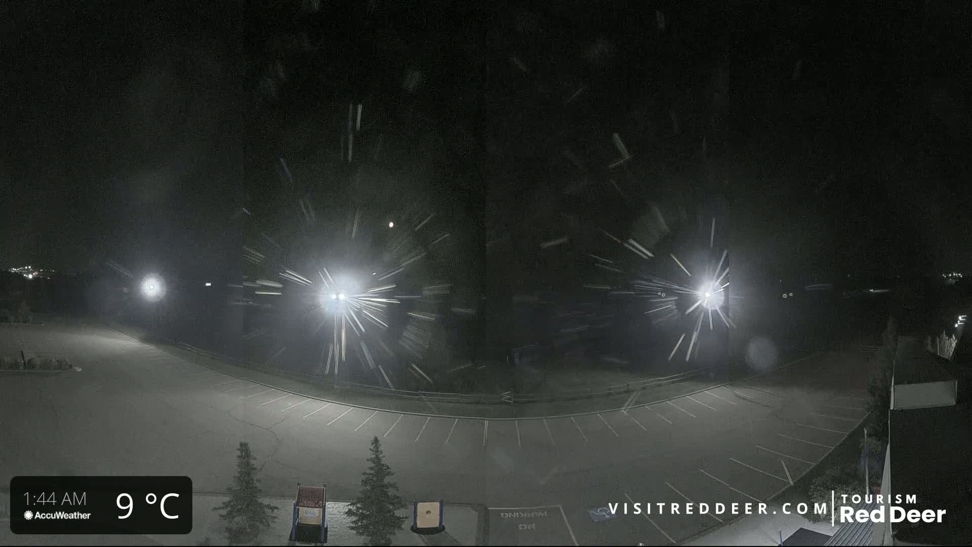 A nighttime view of a snow-covered empty parking lot illuminated by bright lights, with heavy snowfall occurring.