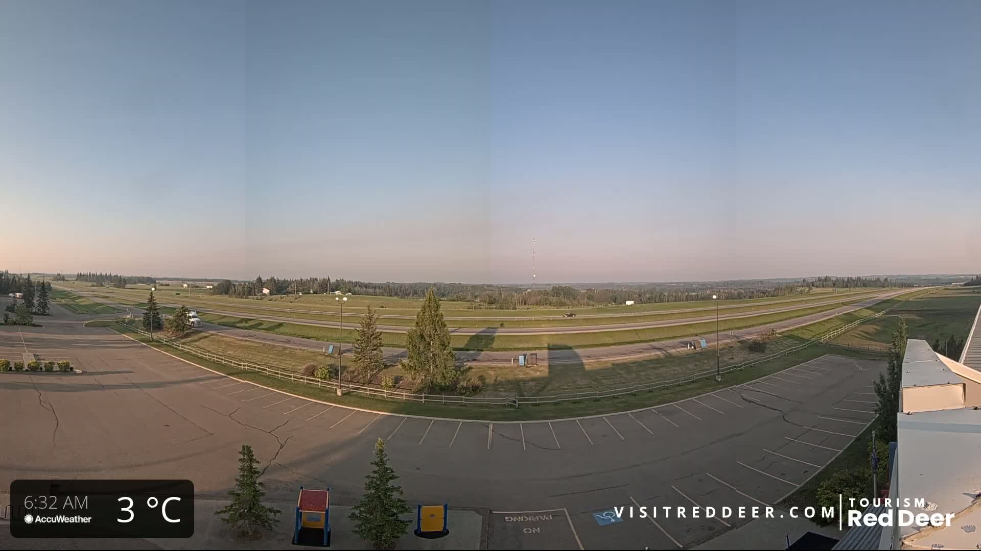 An empty parking lot overlooks a highway and grassy fields under a clear, pale-blue sky at 3°C.