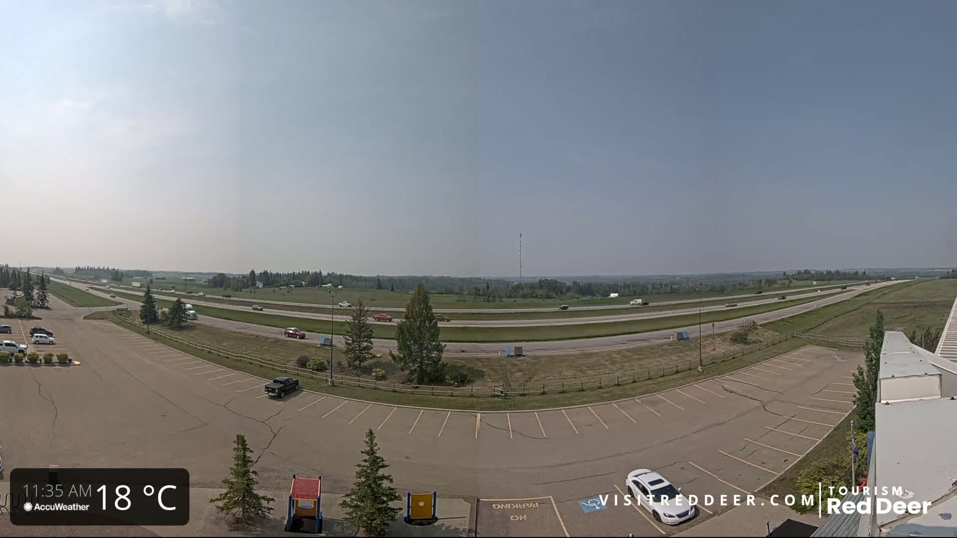 A mostly empty parking lot overlooks a multi-lane highway that stretches into a flat, grassy landscape under a clear, sunny sky.