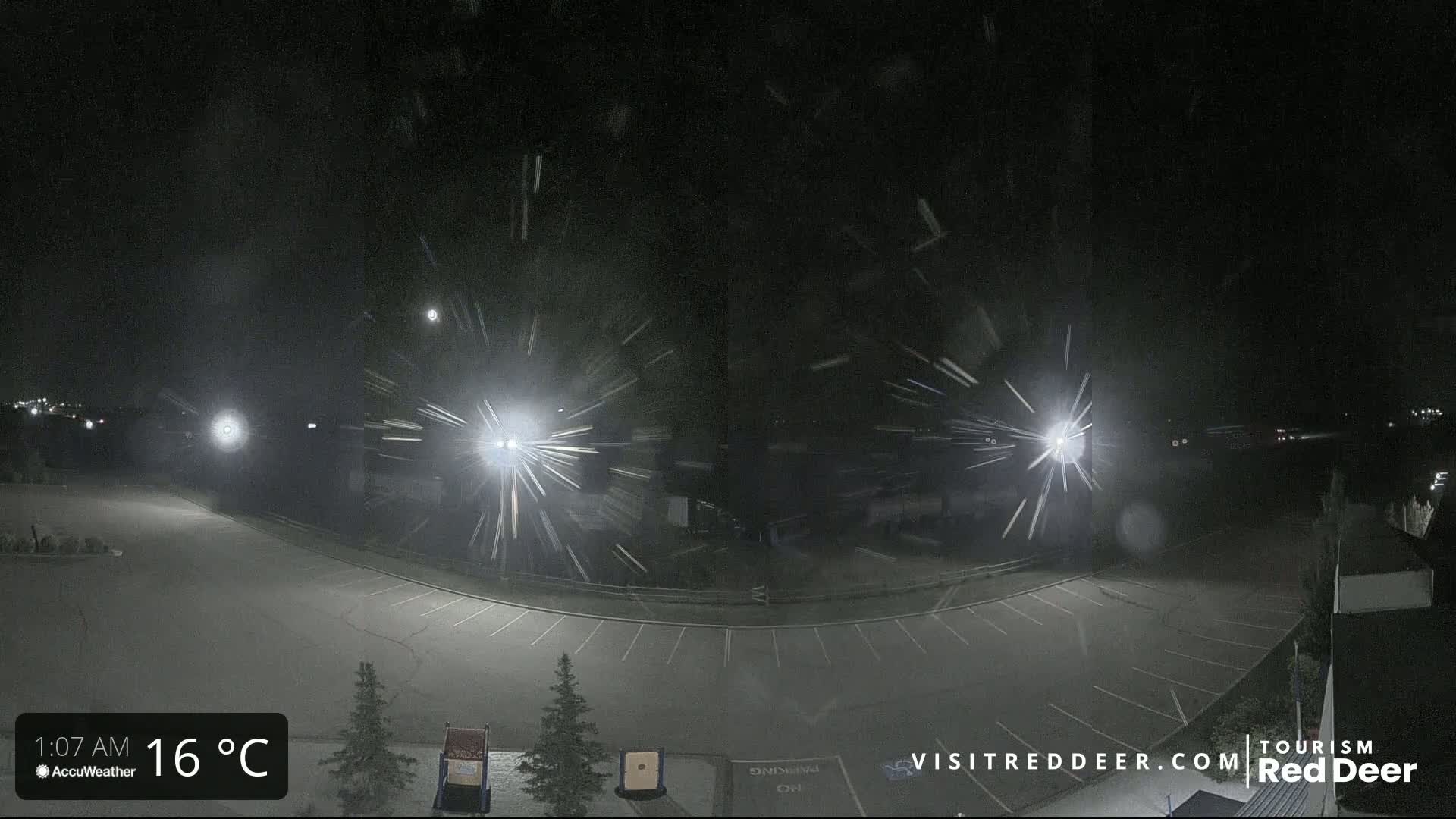 A nighttime, snow-filled scene shows an empty parking lot illuminated by bright lights, with streaks of snow visible in the air.