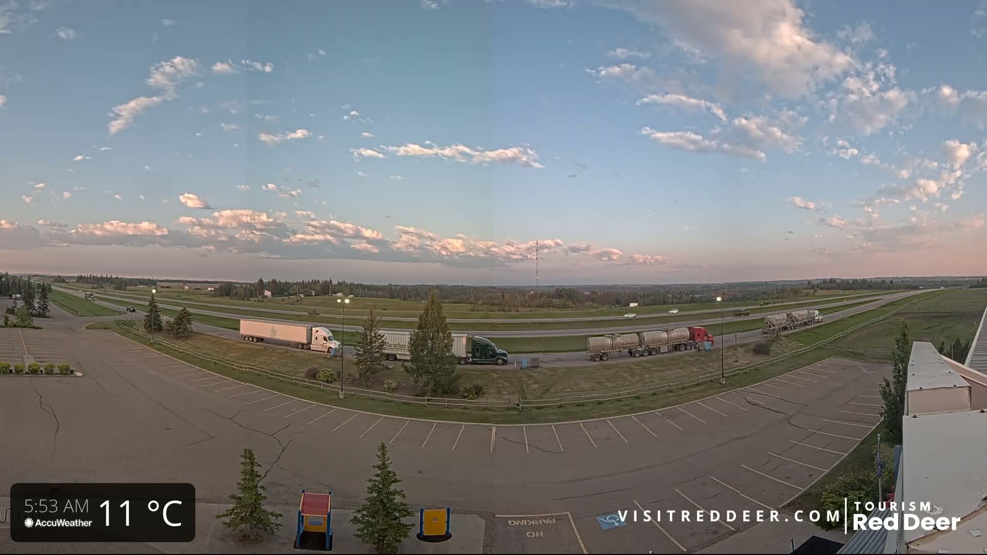 A mostly sunny sky with scattered clouds overlooks a highway with several semi-trucks parked in a rest area adjacent to a large empty parking lot.