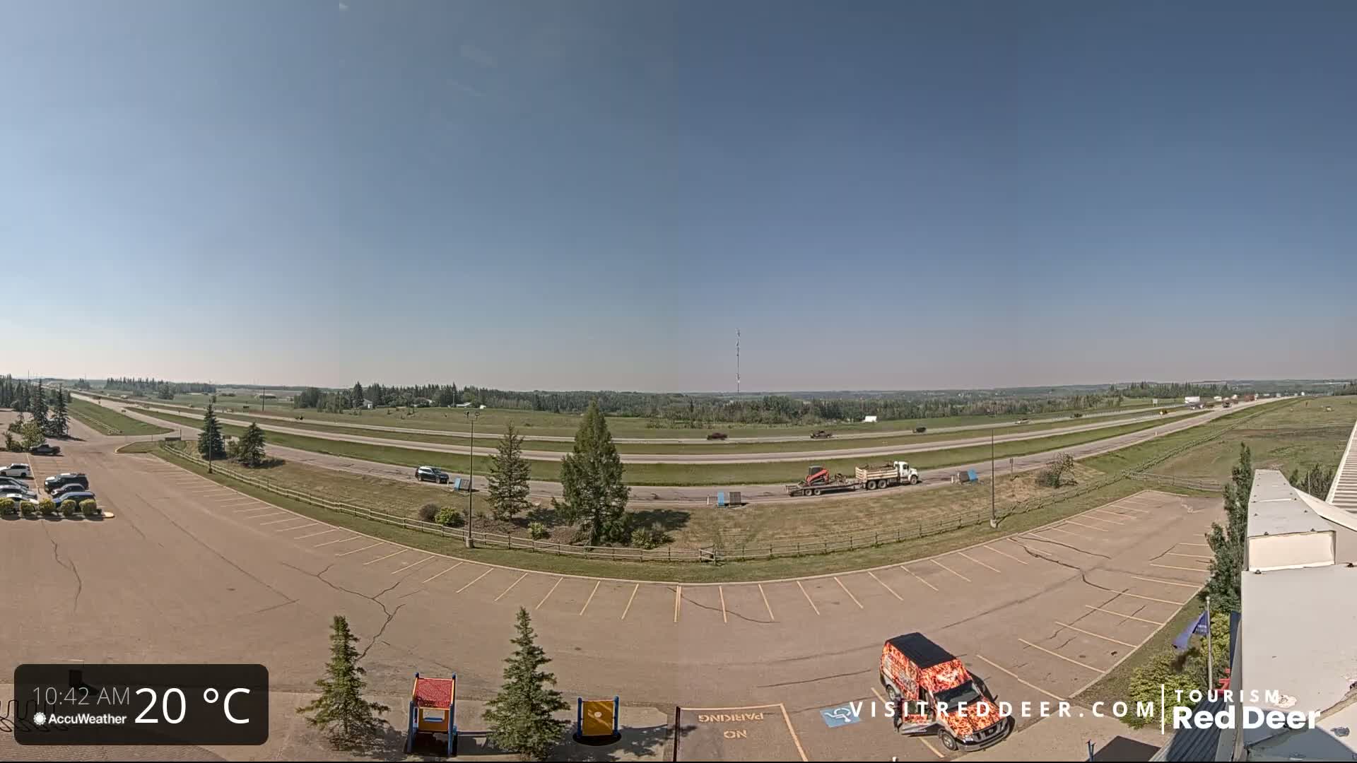 A highway with sparse traffic curves through a flat, grassy landscape under a clear blue sky on a 20°C day, with a parking lot and a small playground visible in the foreground.