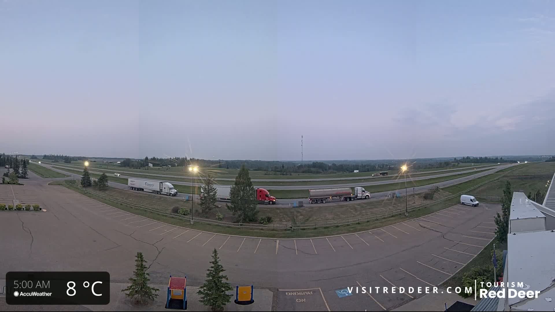 An empty parking lot overlooks a highway with several semi-trucks and a van, under a clear, twilight sky at 8°C.
