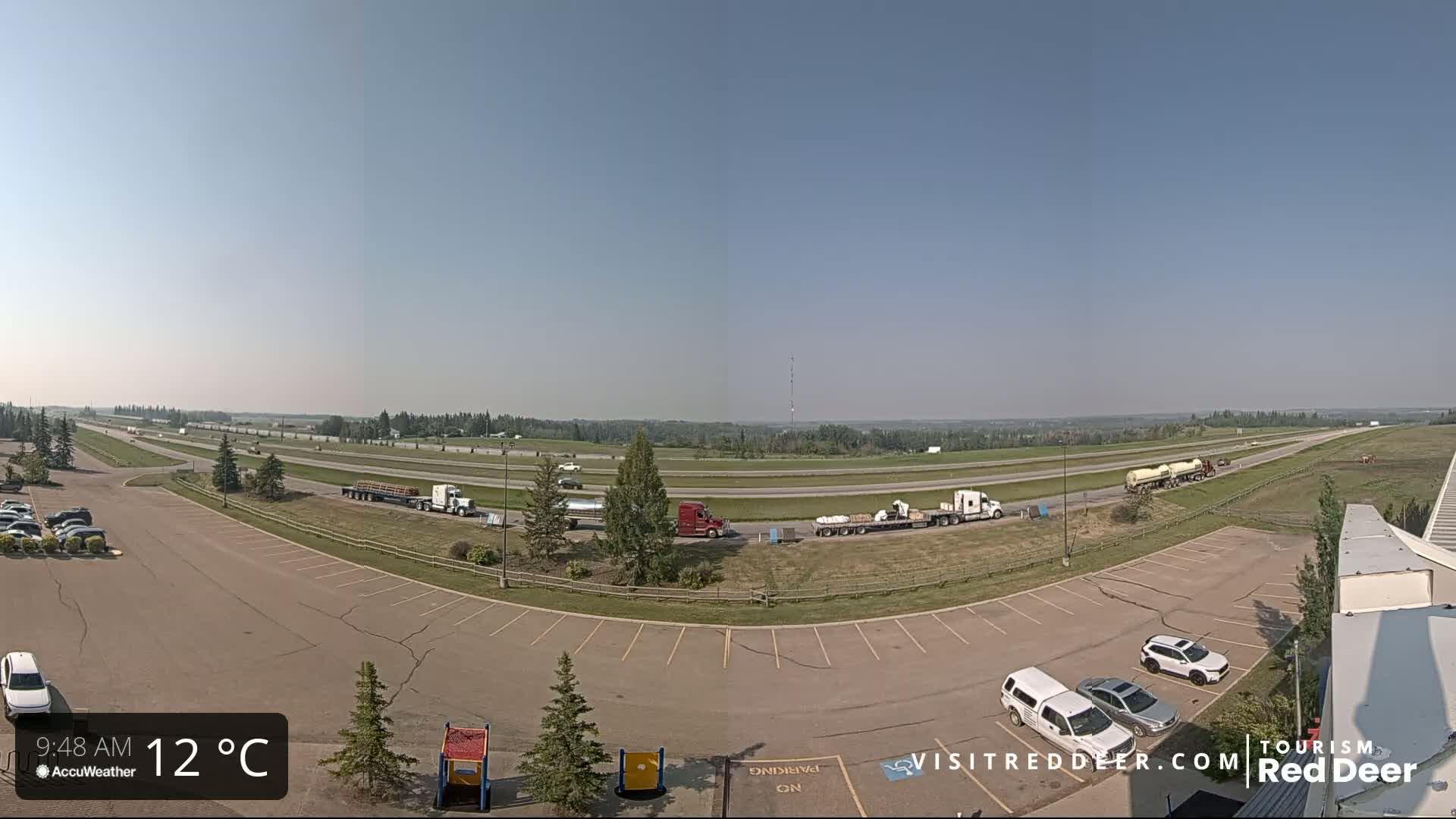 A highway with several semi-trucks and cars is visible from a high vantage point on a clear, sunny day, with a parking lot and some small playground equipment in the foreground.