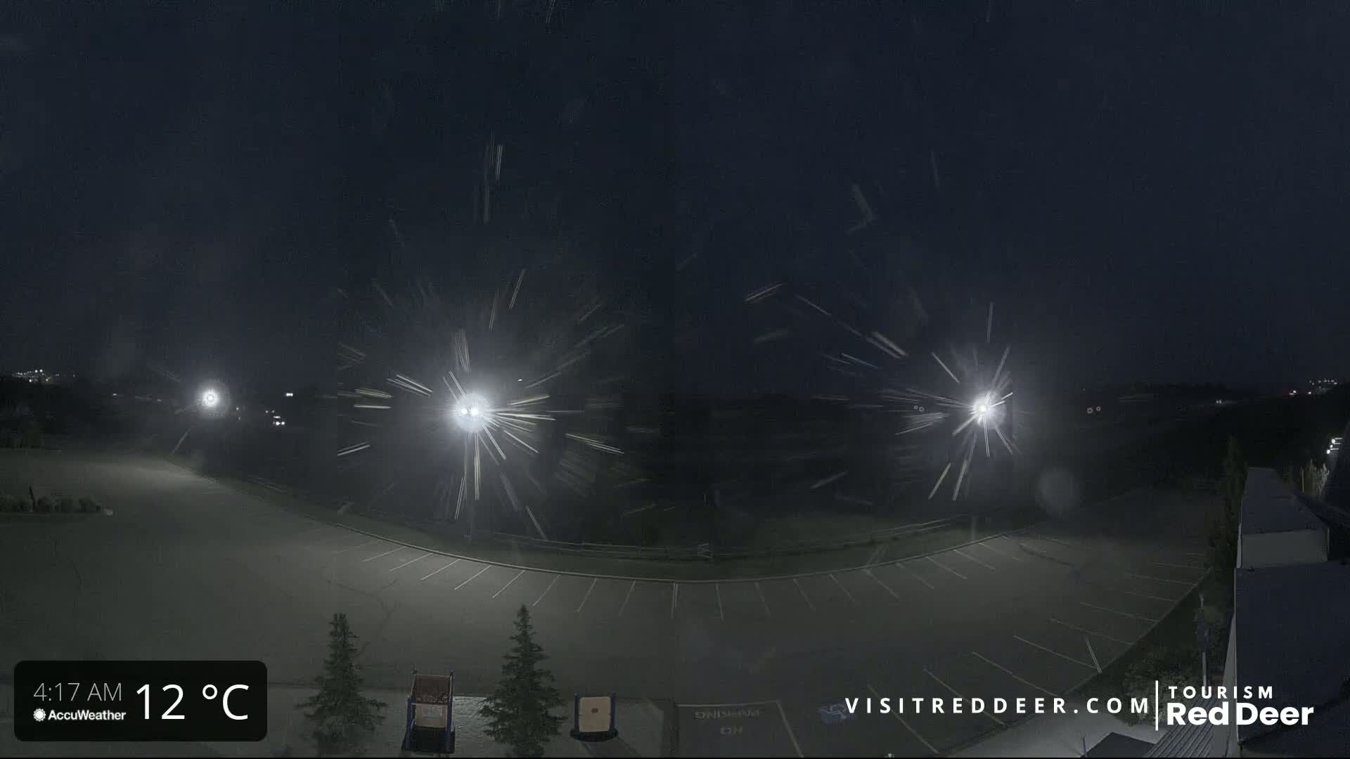 A nighttime, snow-filled scene shows an empty parking lot illuminated by bright lights, with a few cars visible in the distance.