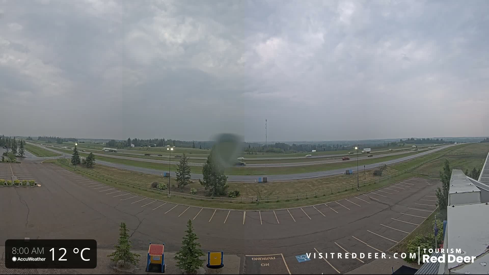 A mostly cloudy day shows a highway with sparse traffic, an empty parking lot, and some small trees under an overcast sky.