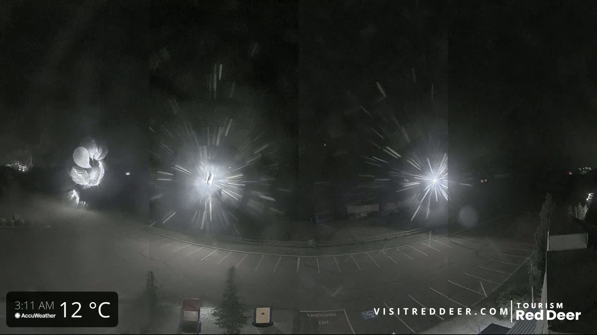 A nighttime, snowy scene shows a mostly empty parking lot illuminated by bright lights, with snow appearing as streaks in the image.