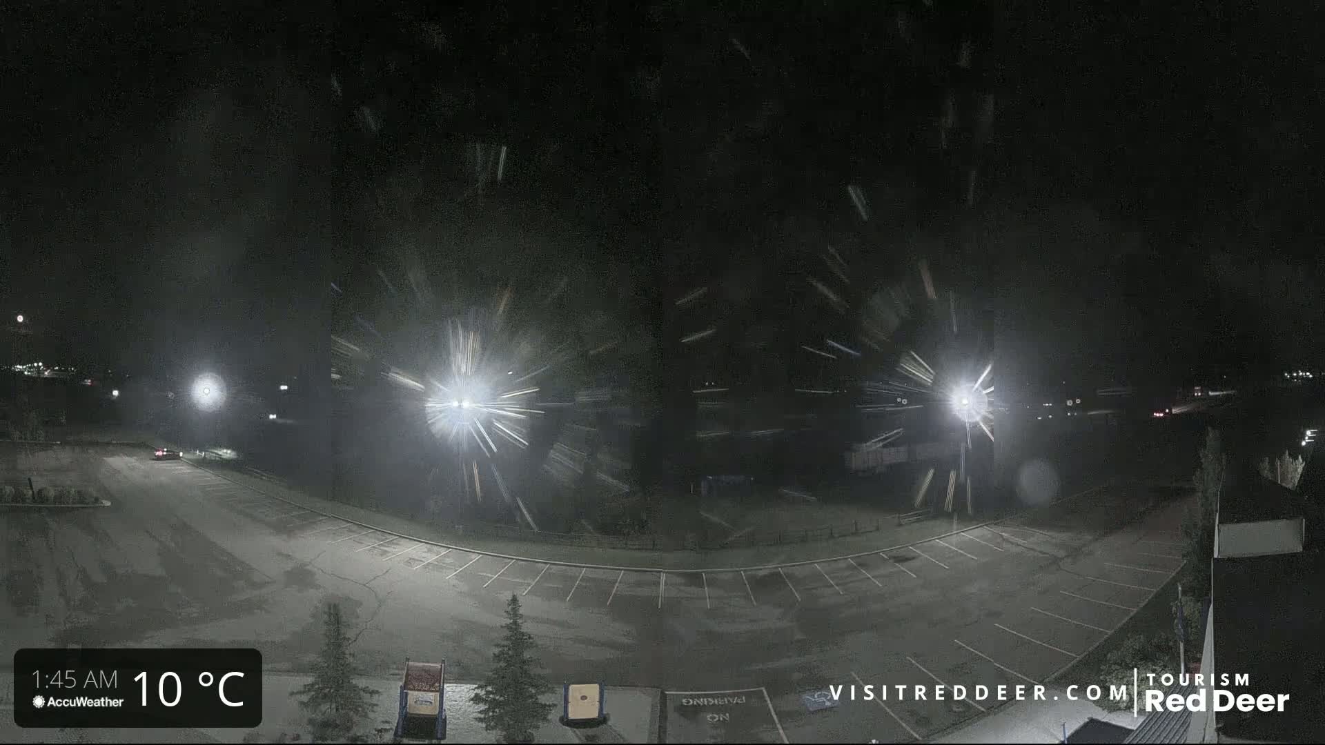 A nighttime view of a snow-covered parking lot with several cars and streetlights, under heavy snowfall.