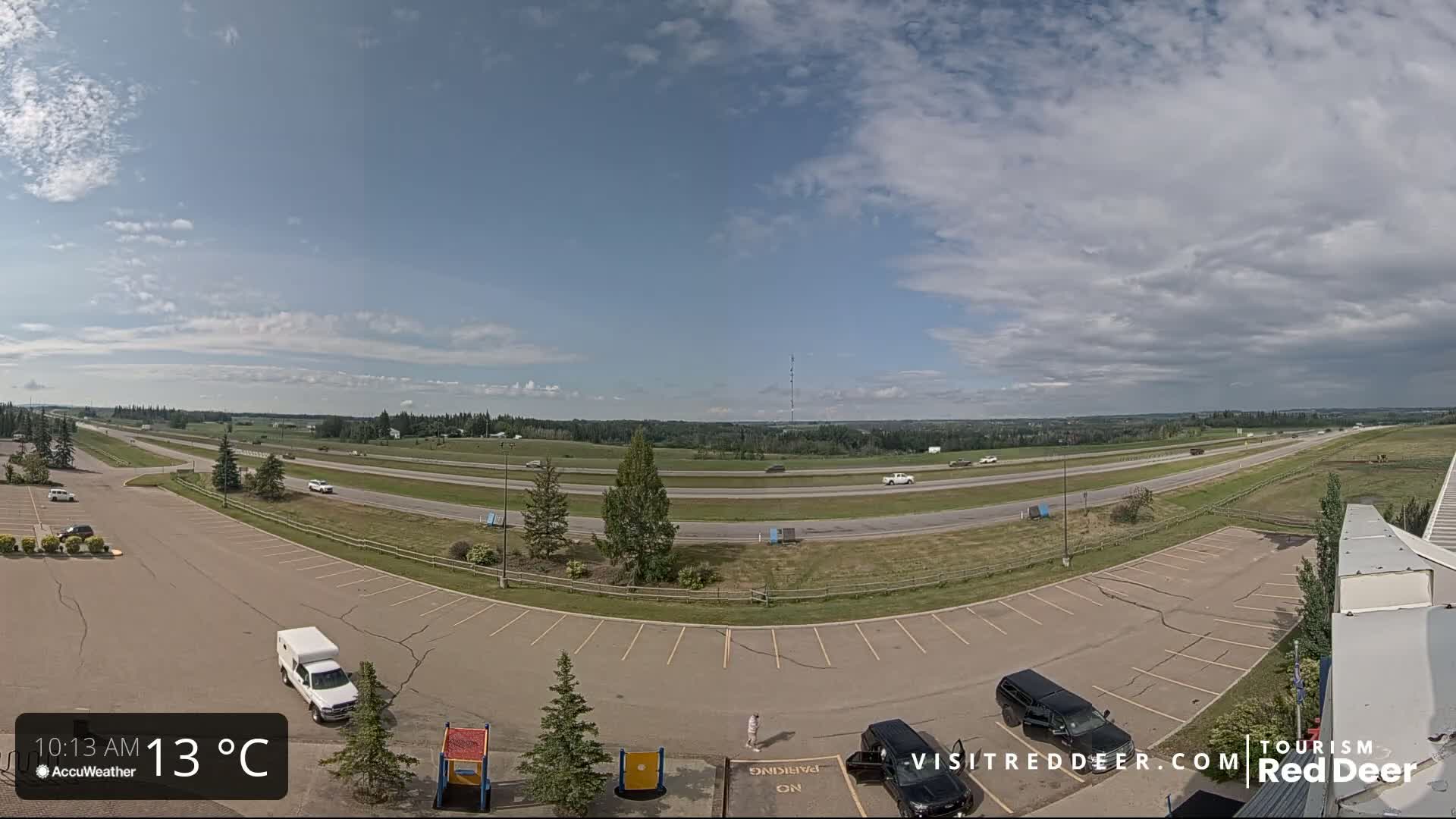 A partly cloudy sky overlooks a highway with sparse traffic and an empty parking lot containing a few vehicles, some playground equipment, and a small amount of greenery.