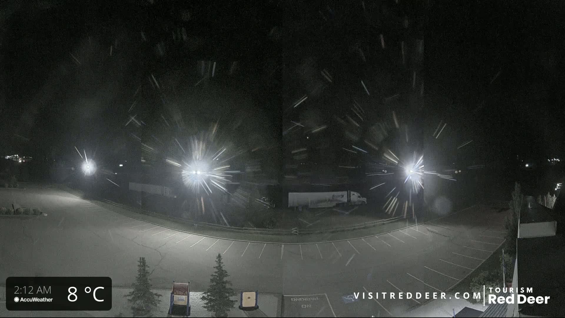 A nighttime, snowy scene shows an empty parking lot illuminated by bright lights, with some vehicles visible in the background.
