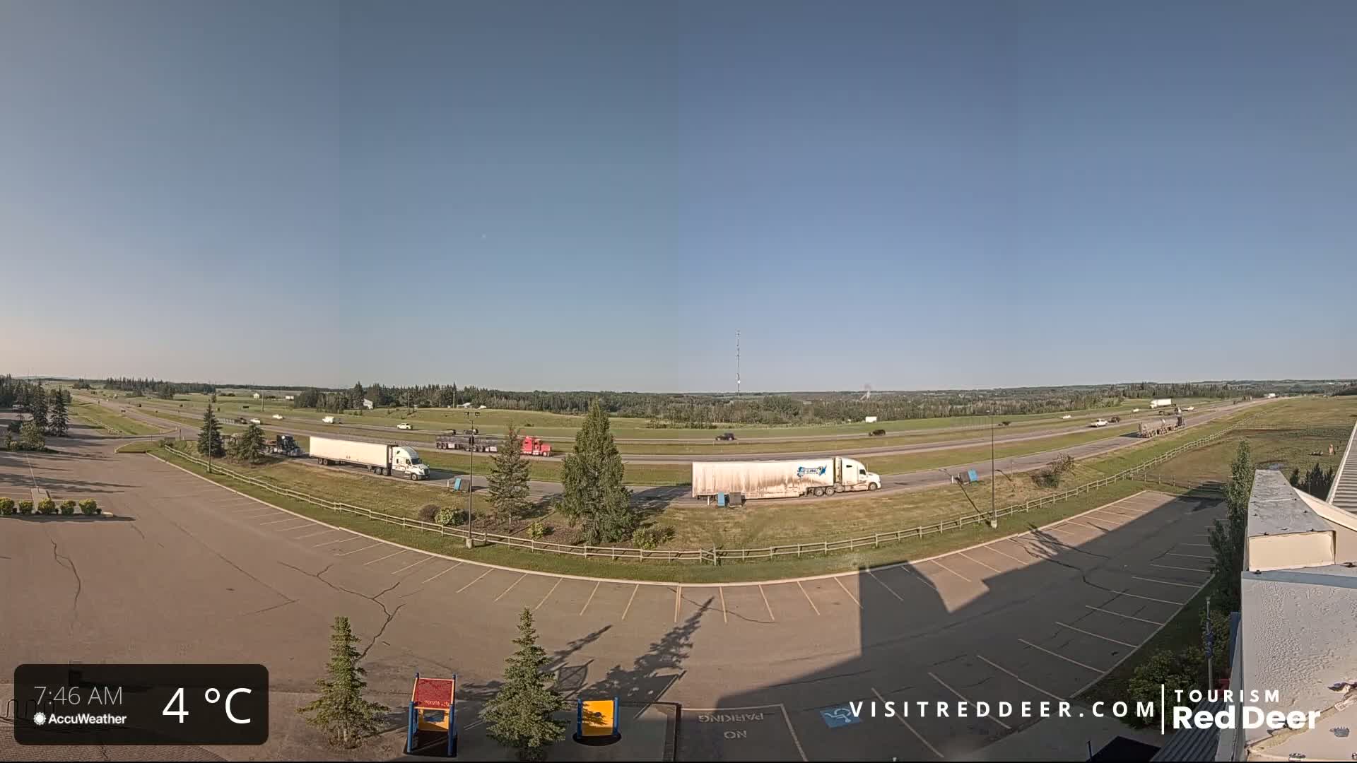 A mostly empty parking lot overlooks a highway with several semi-trucks and cars driving under a clear, sunny sky.