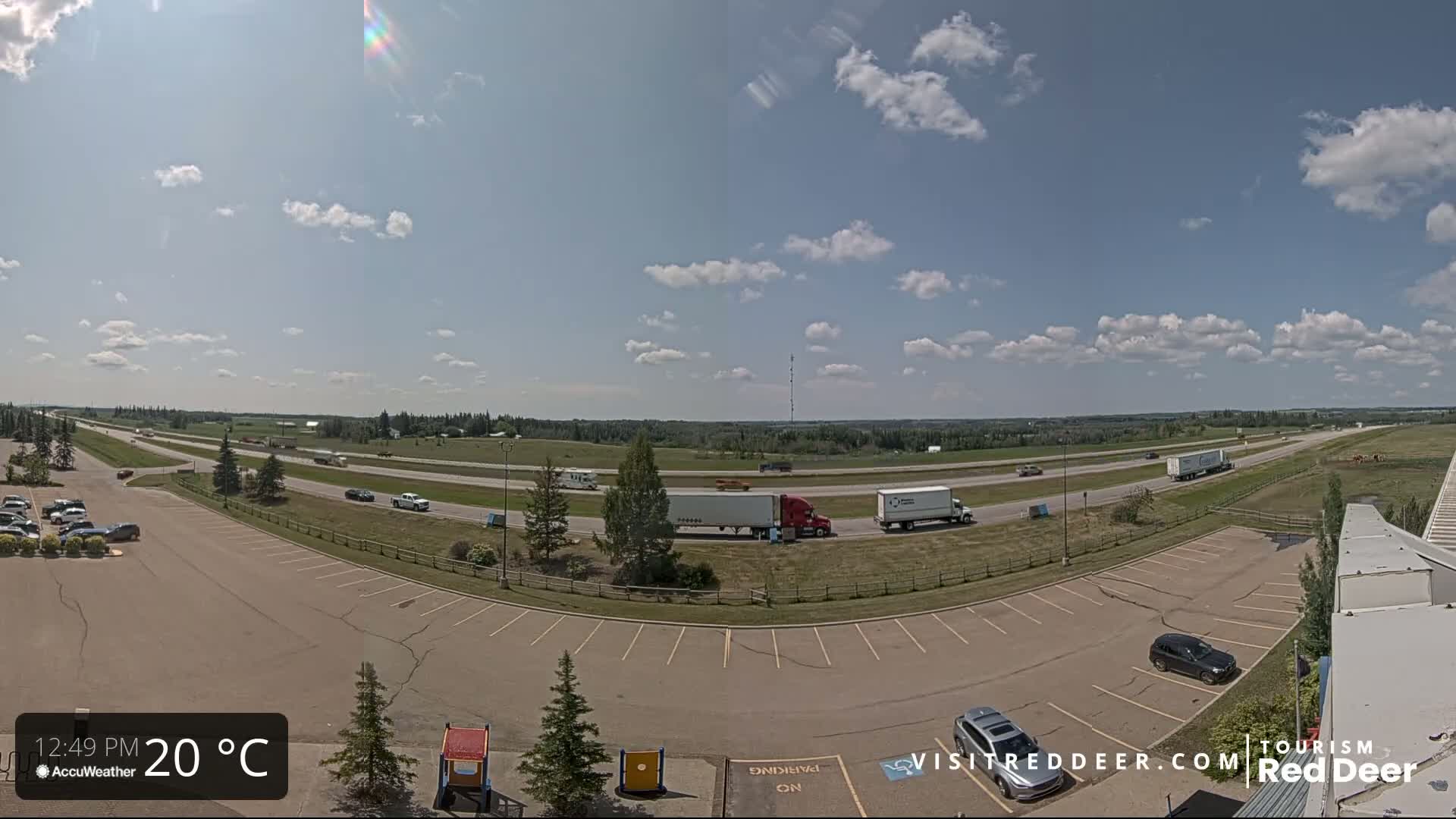A mostly sunny day shows a highway with several vehicles, including semi-trucks, and a large empty parking lot next to it.