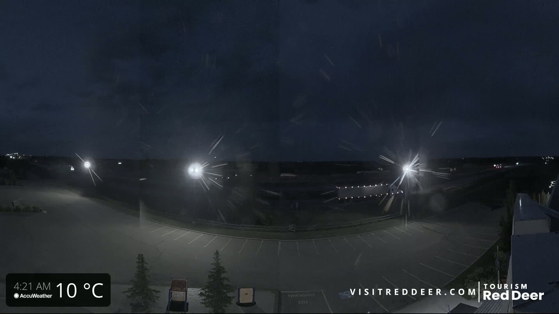 A nighttime view of an empty parking lot with light snow falling under a dark sky, illuminated by several bright lights.