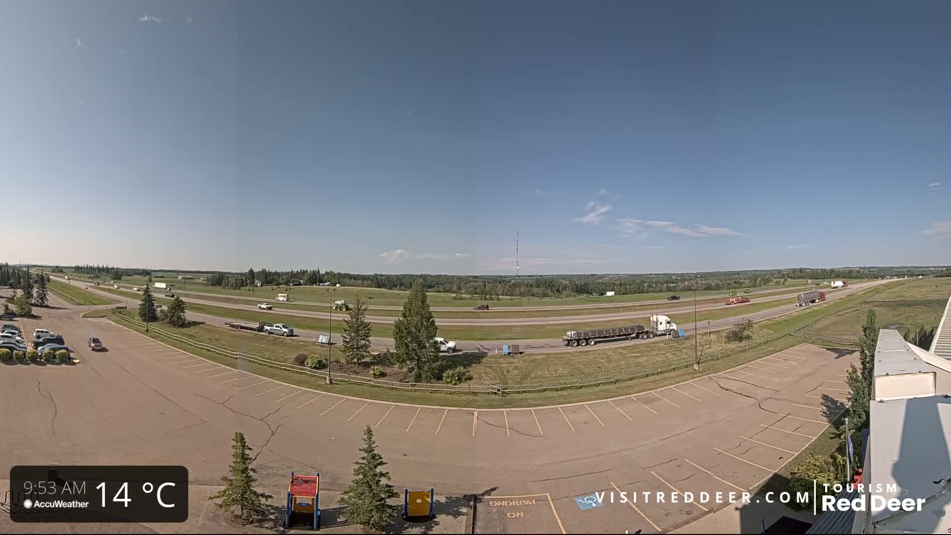 A mostly empty parking lot overlooks a highway with several vehicles, under a clear blue sky on a 14°C day.