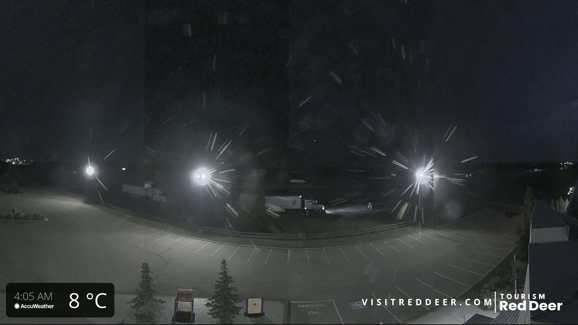 A nighttime view of a mostly empty parking lot with a semi-truck in the distance during a light snow flurry.