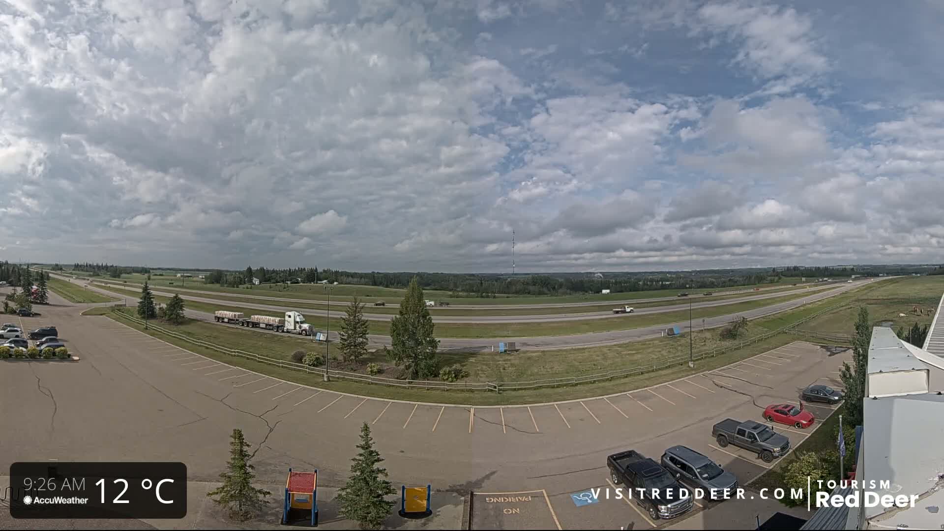 A partly cloudy day reveals a highway with several vehicles, including a semi-truck, next to a parking lot with cars and a grassy area beyond.
