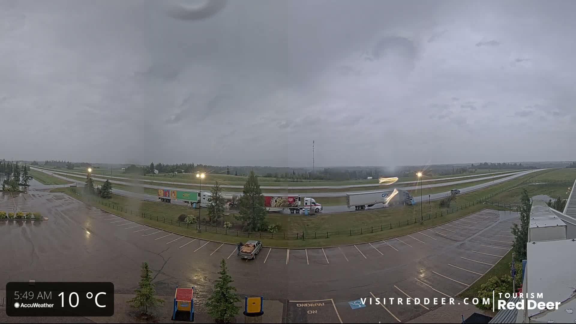A wet parking lot and highway are visible under an overcast sky, with several semi-trucks parked and driving along the highway.