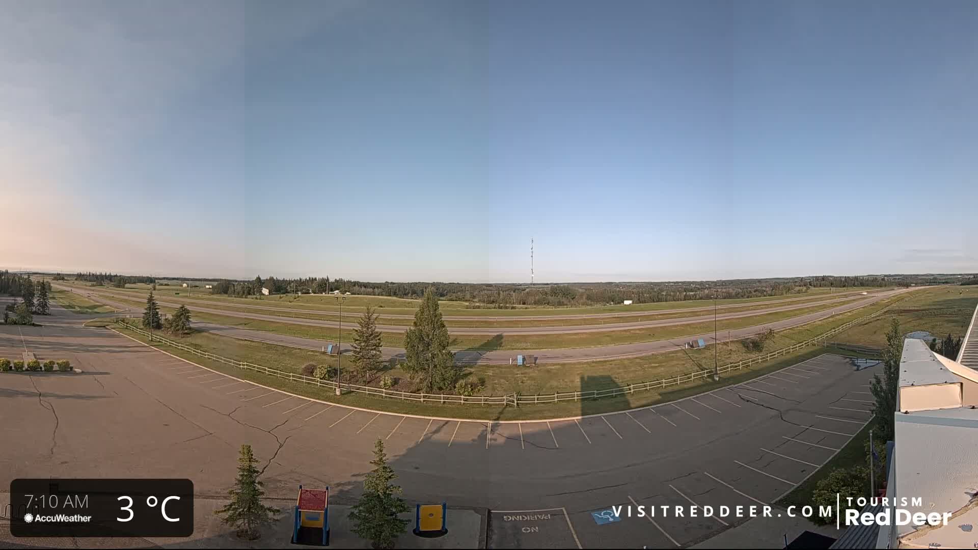 An empty parking lot overlooks a multi-lane highway stretching across a flat landscape under a clear, bright sky.