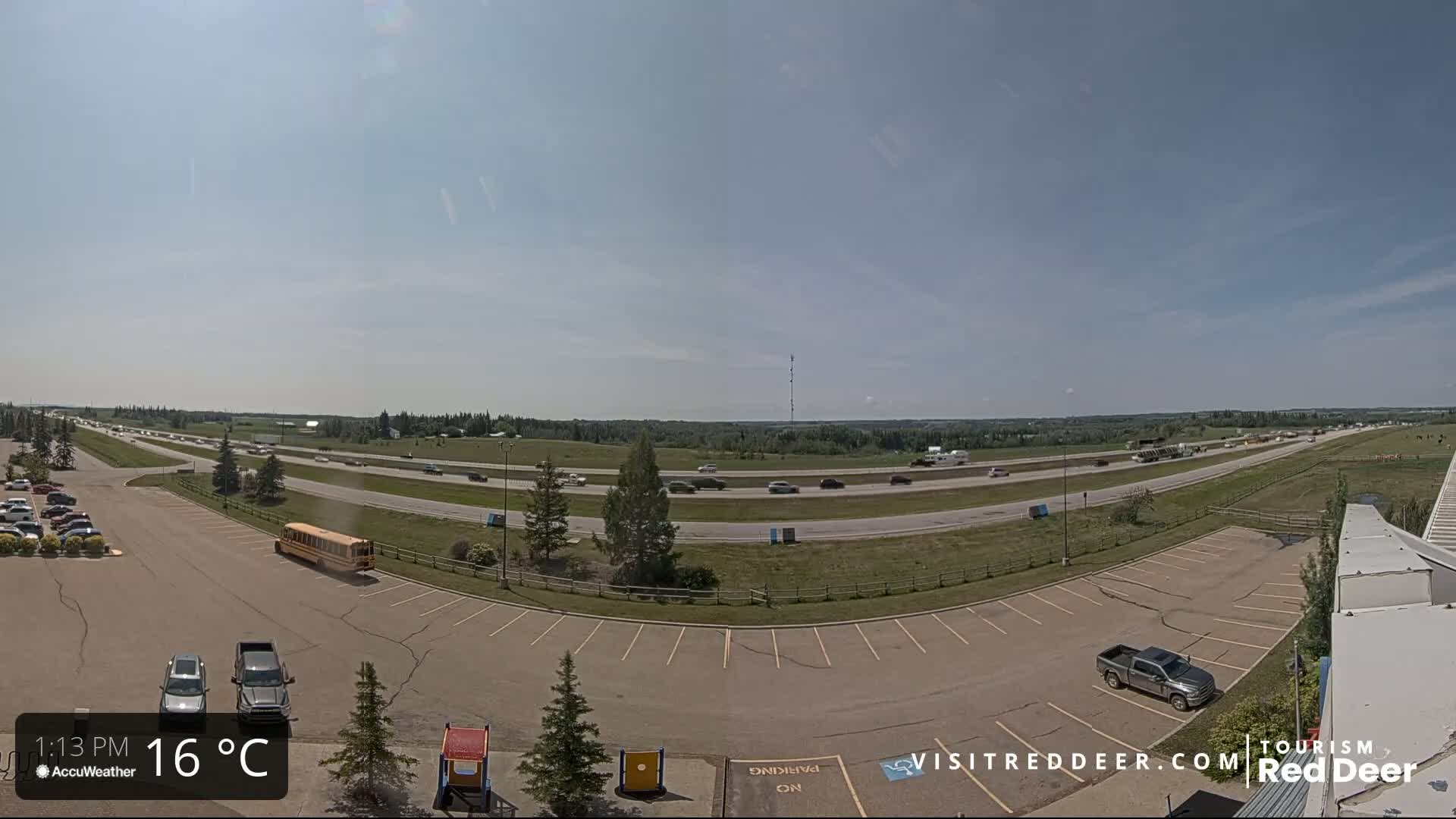 A highway with moderate traffic is visible on a sunny day, with a parking lot and a few vehicles in the foreground.