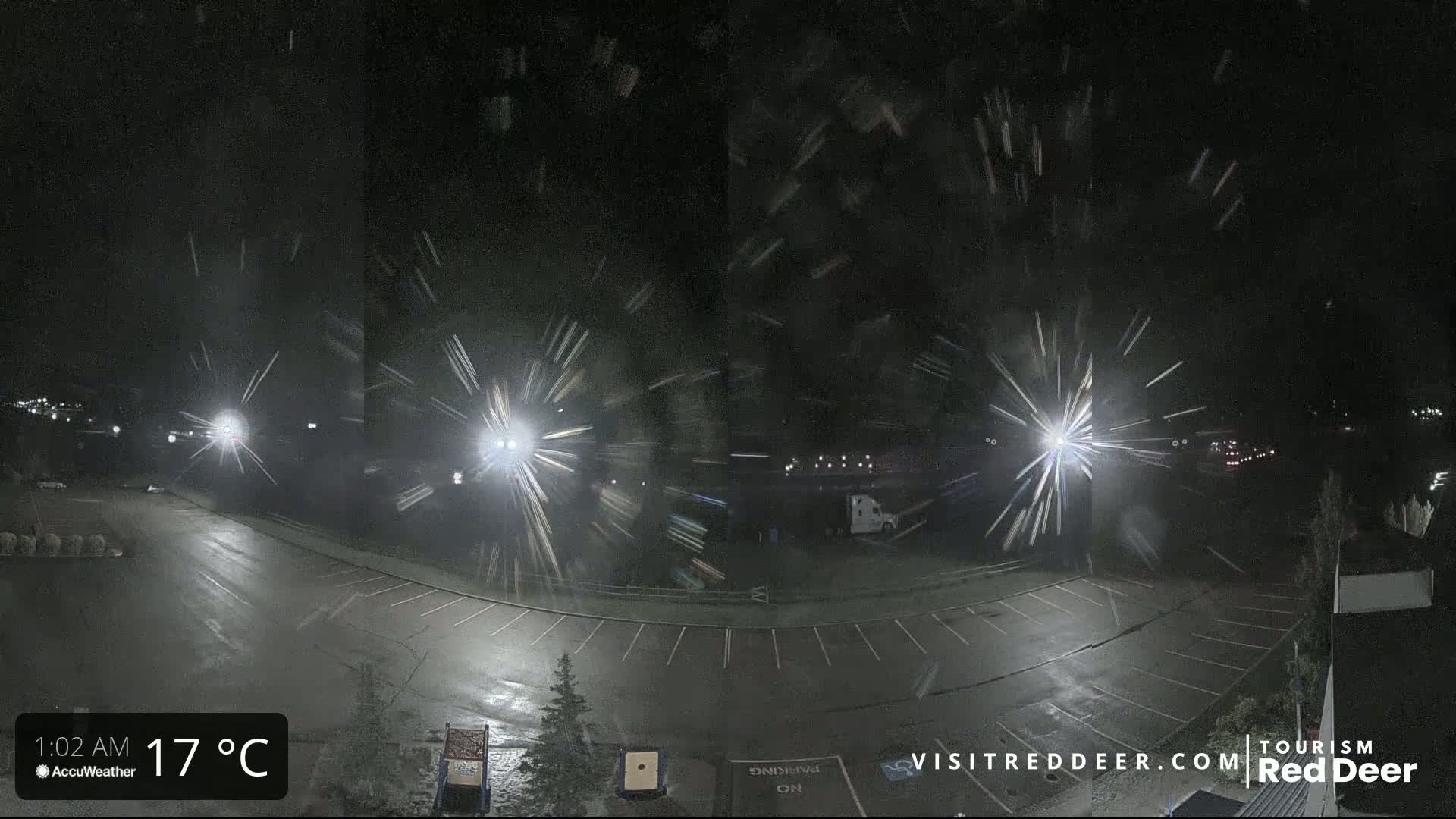 A nighttime view of a mostly empty parking lot with light snow falling, illuminated by several bright lights.