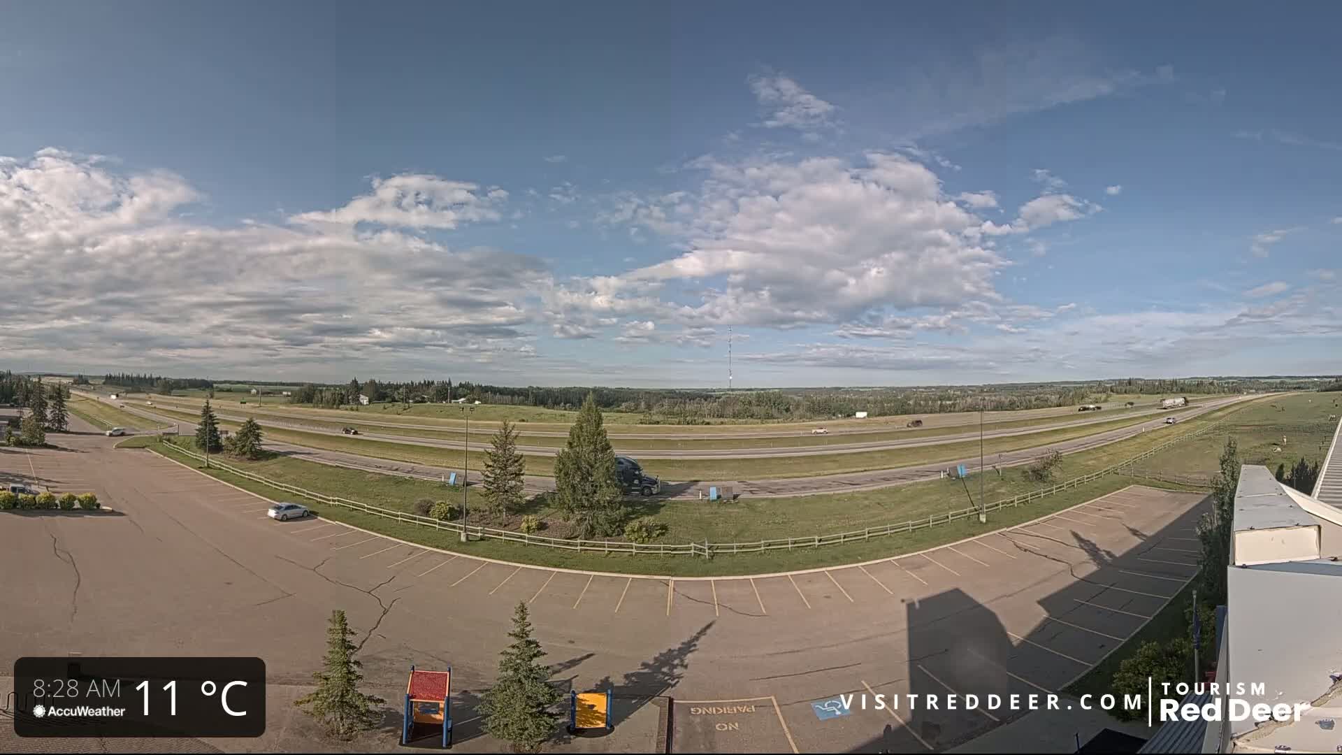A partly cloudy sky overlooks a highway with several vehicles, an empty parking lot, and some sparse vegetation.