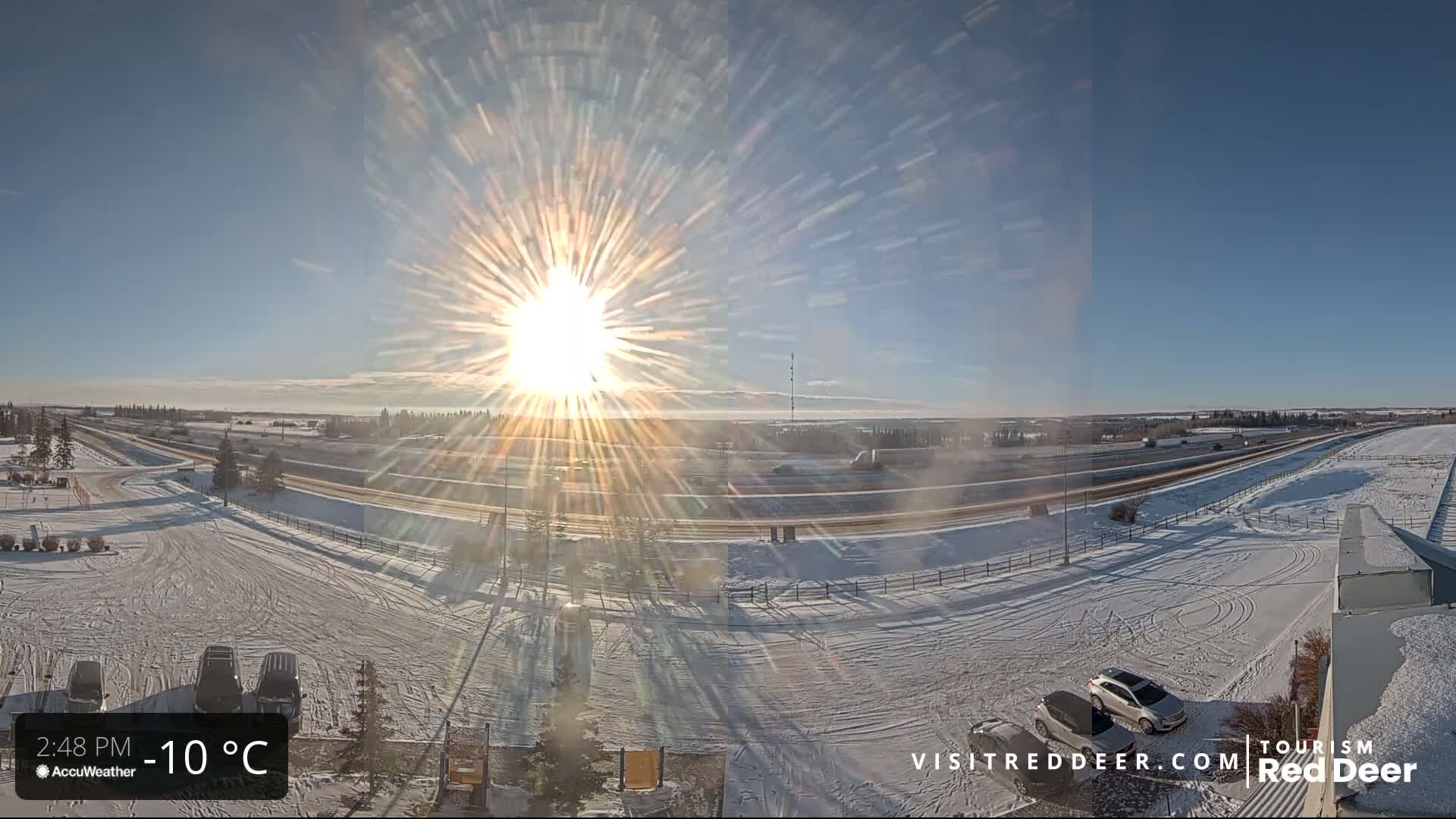 A sunny and clear winter day features a brilliant sun casting long shadows across snow-covered roads, fields, and a parking area with a few vehicles.
