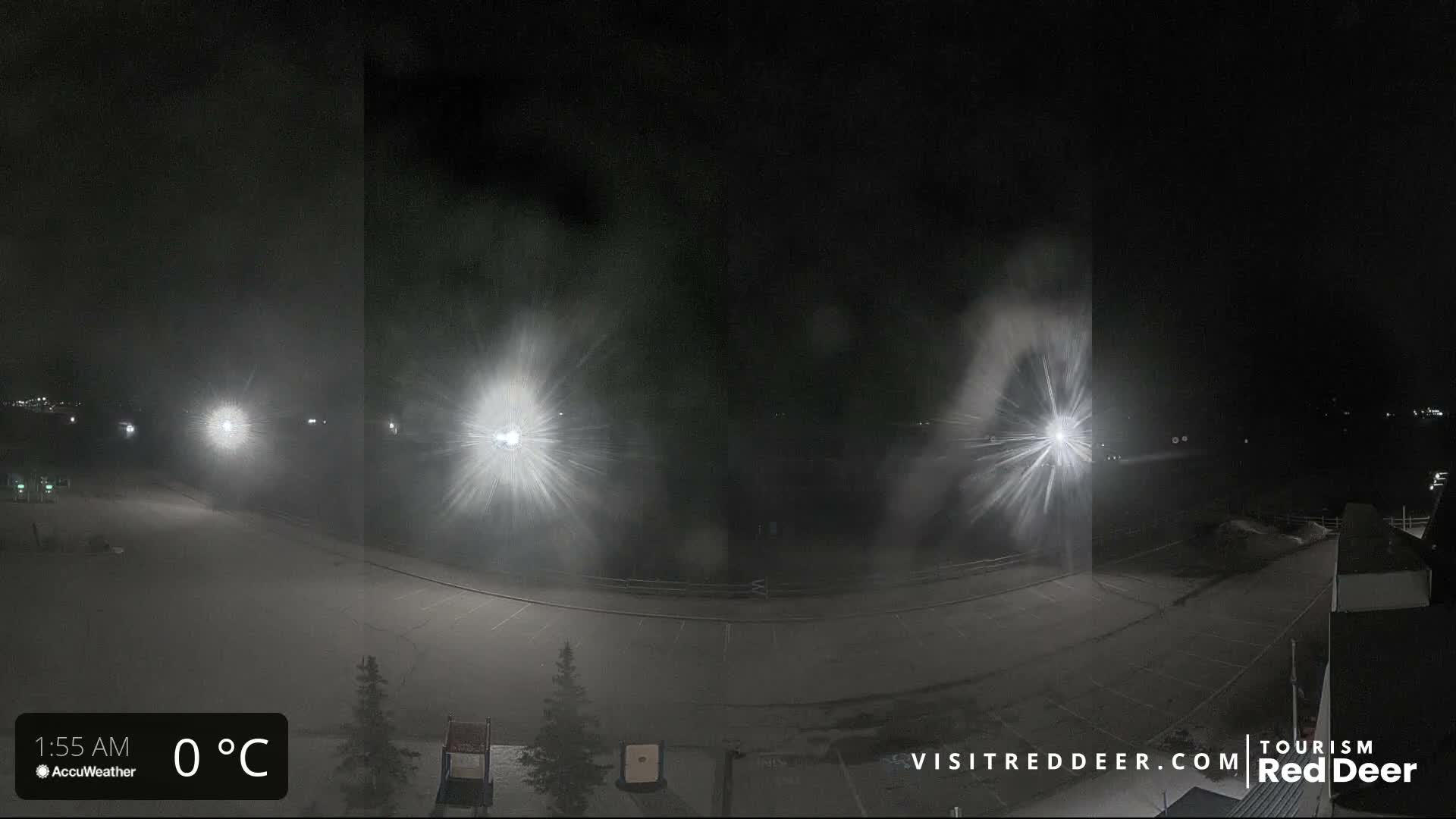 A wide nighttime view shows a snow-covered landscape illuminated by distant bright lights that create pronounced starbursts through heavy falling snow.