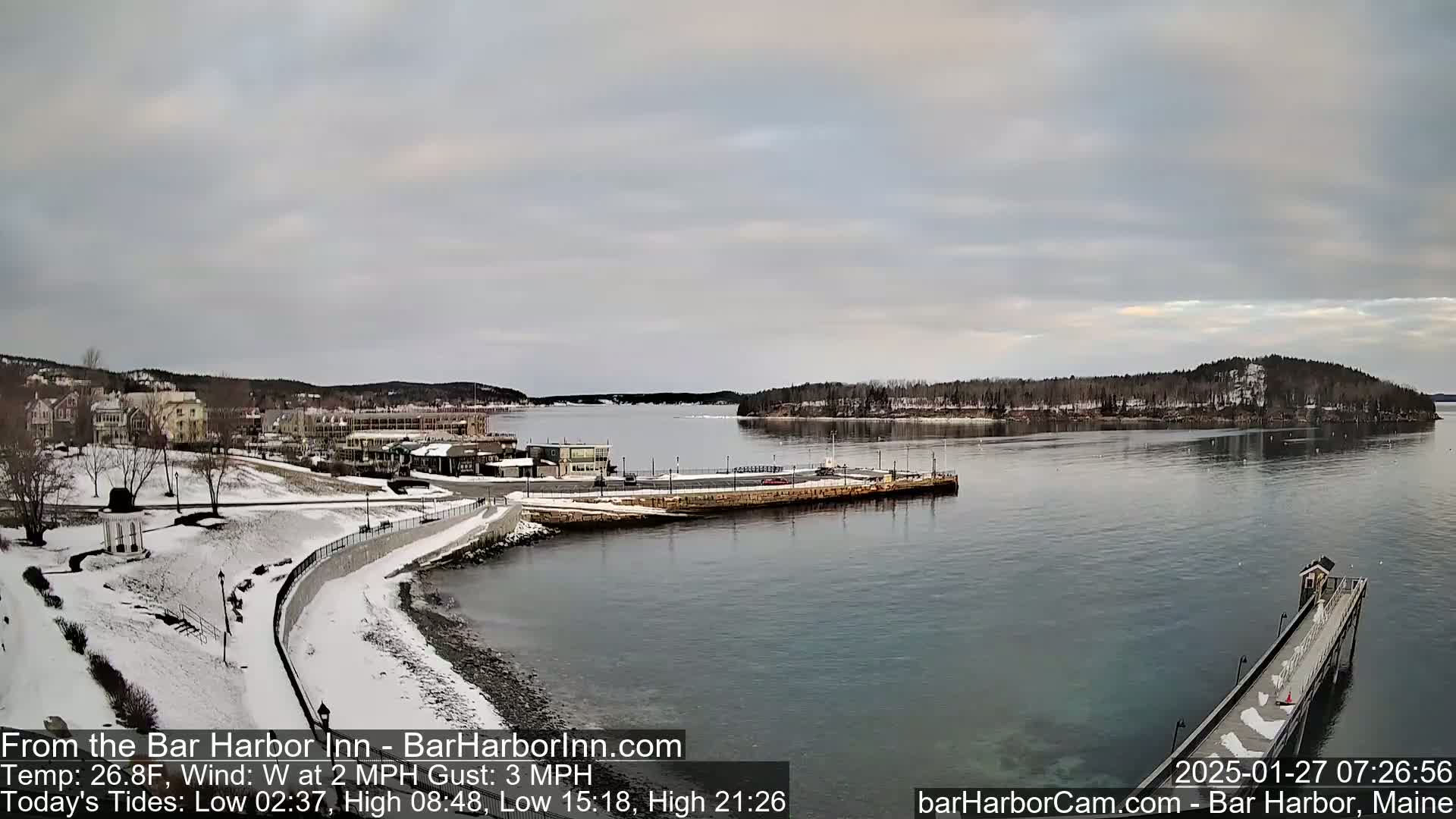 A snow-covered waterfront town is seen on an overcast day, with a calm body of water and a long pier extending into the bay.