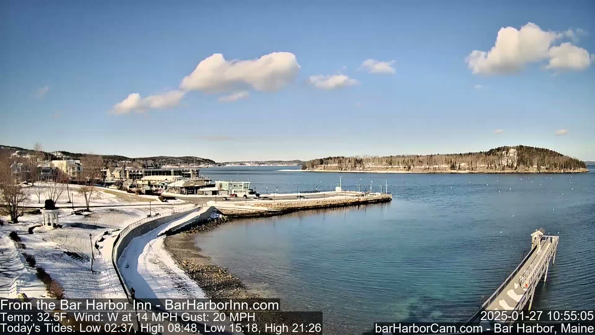 A partly cloudy, cold day with a view of a snow-dusted harbor town, a calm body of water, and a long pier extending into the sea.